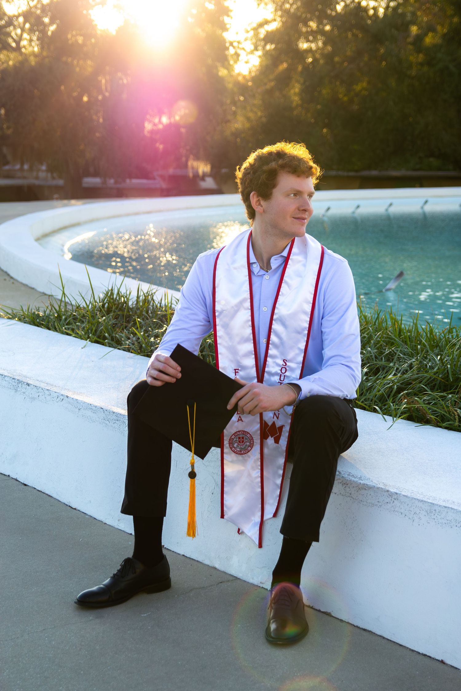Graduate in white shirt and red suspenders sits by pool at sunset holding diploma with tassel and honor cords.