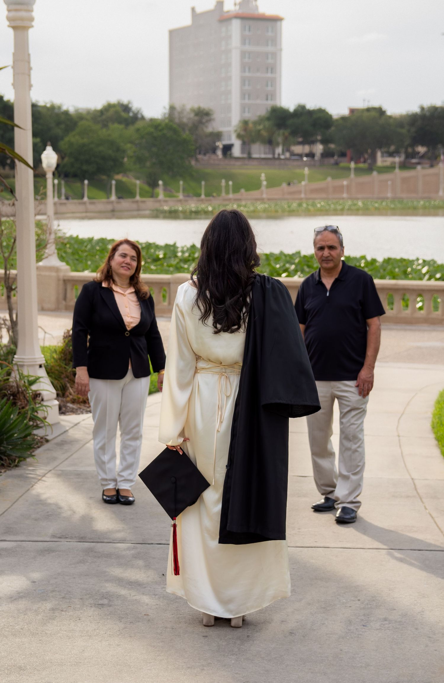 Graduate in cap and gown poses with proud parents outdoors near a scenic waterfront on graduation day.