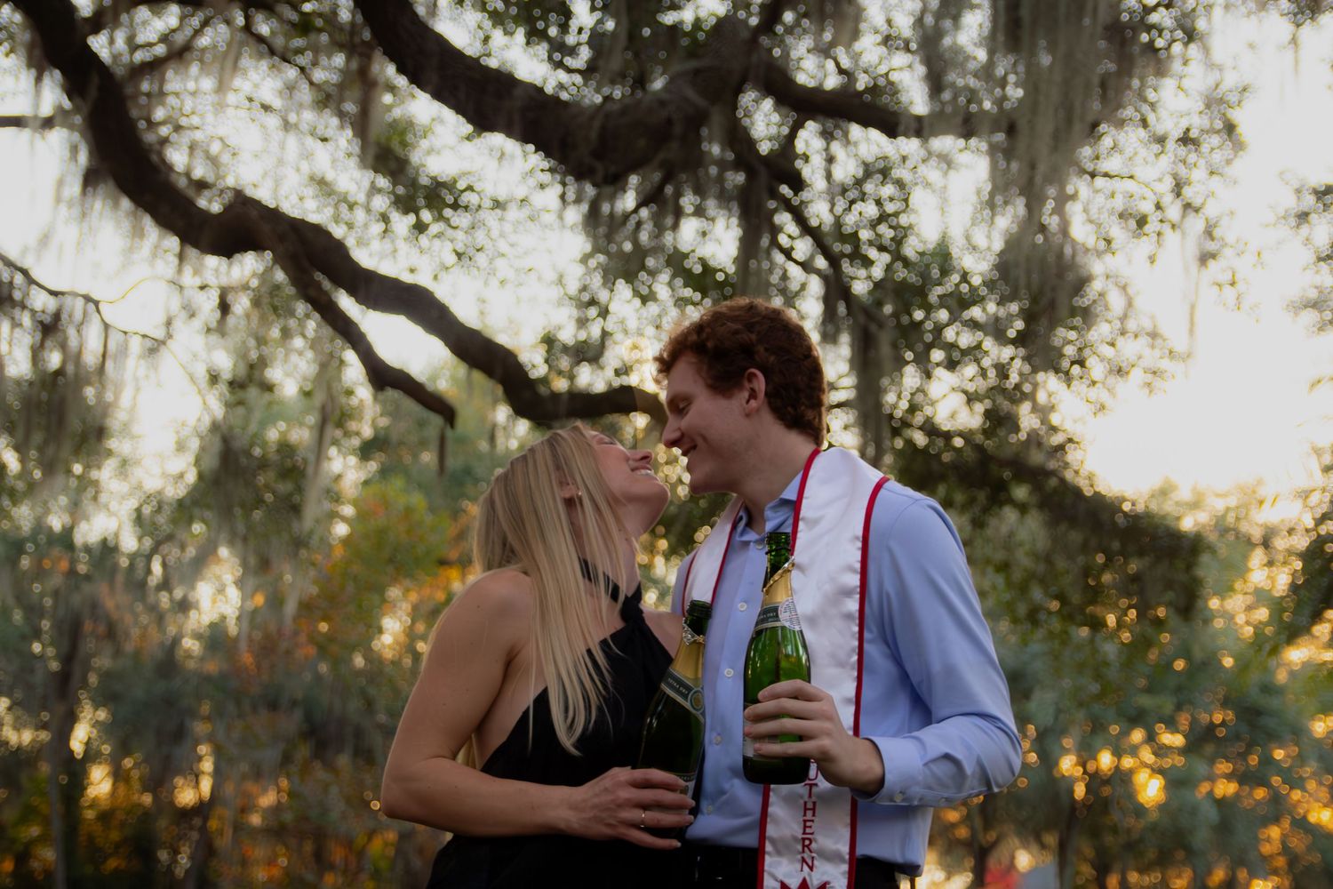 A couple shares a romantic moment with drinks under Spanish moss at sunset in an outdoor setting.
