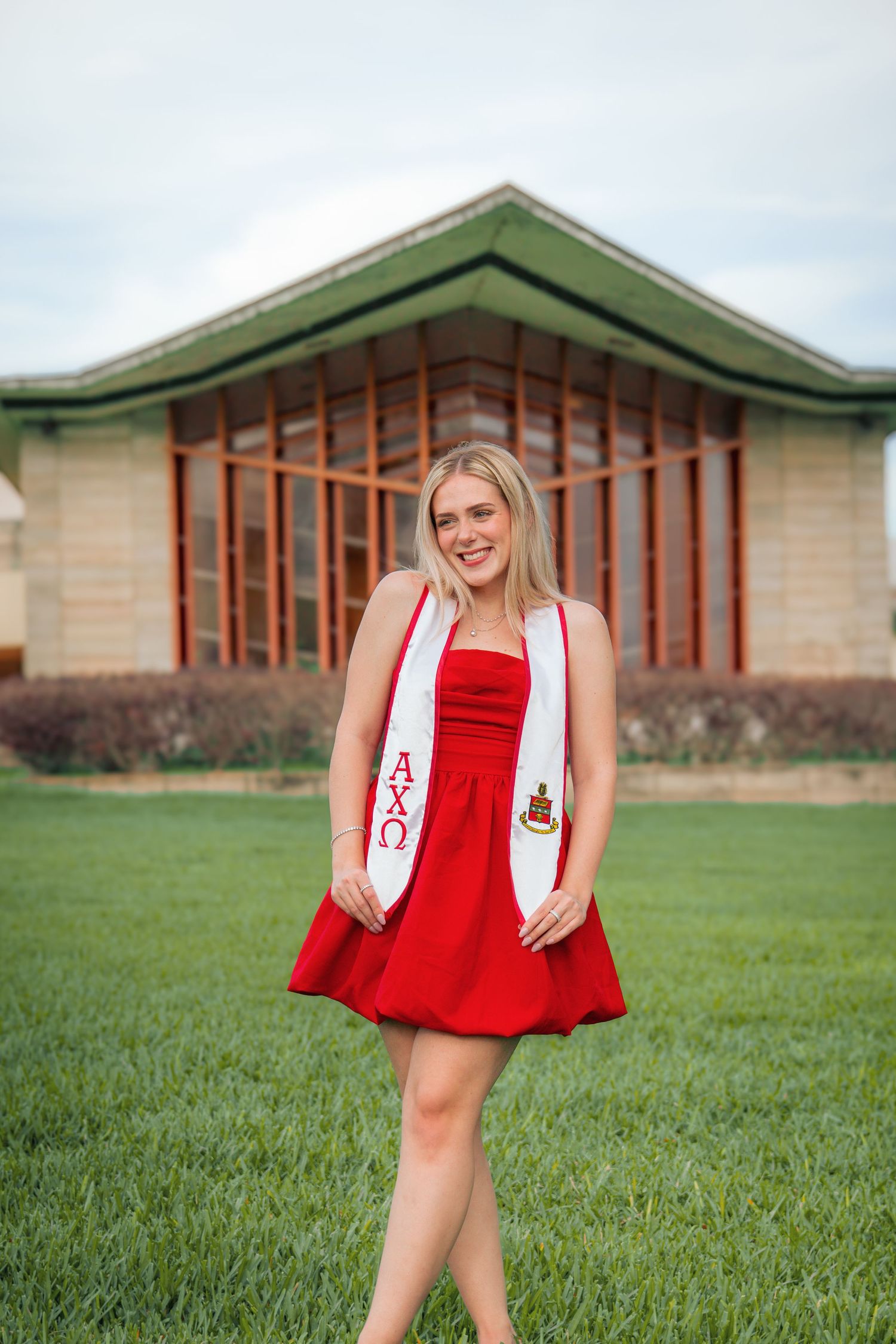 A person in a red dress twirls playfully on a green lawn in front of a brick building.