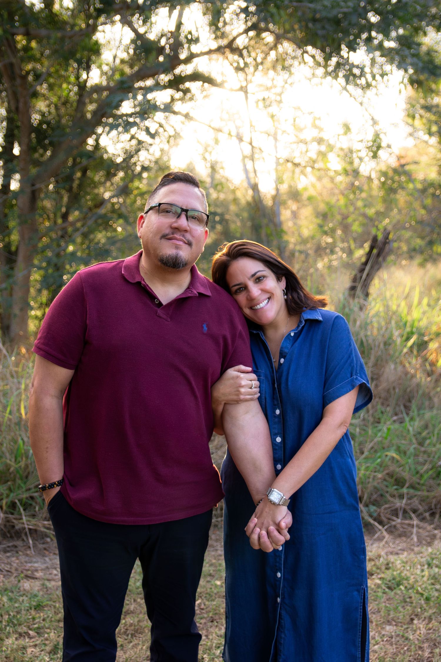 A couple poses together in a natural outdoor setting with golden sunlight filtering through trees in the background.
