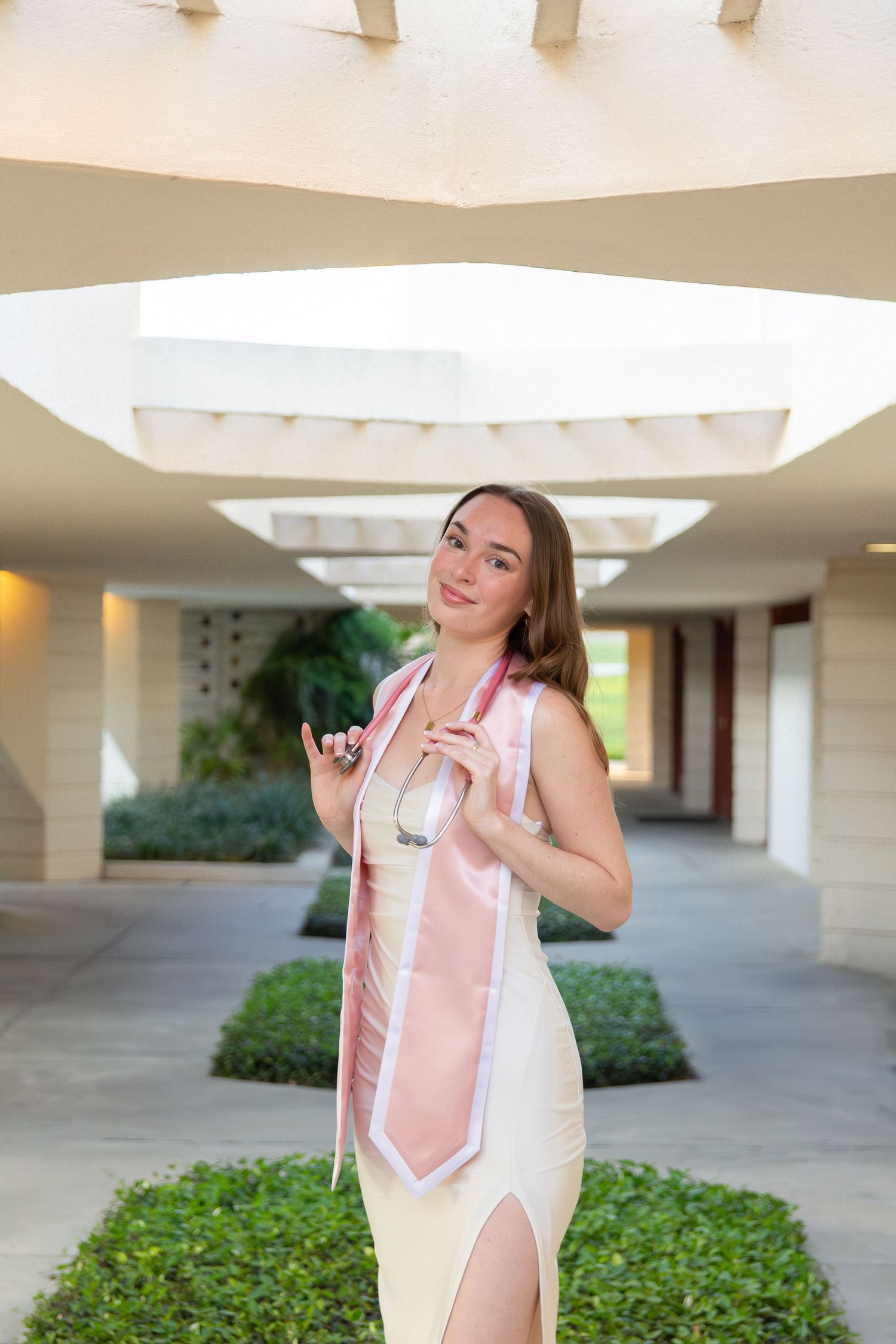 Graduate in white dress posing in modern campus architecture hallway.