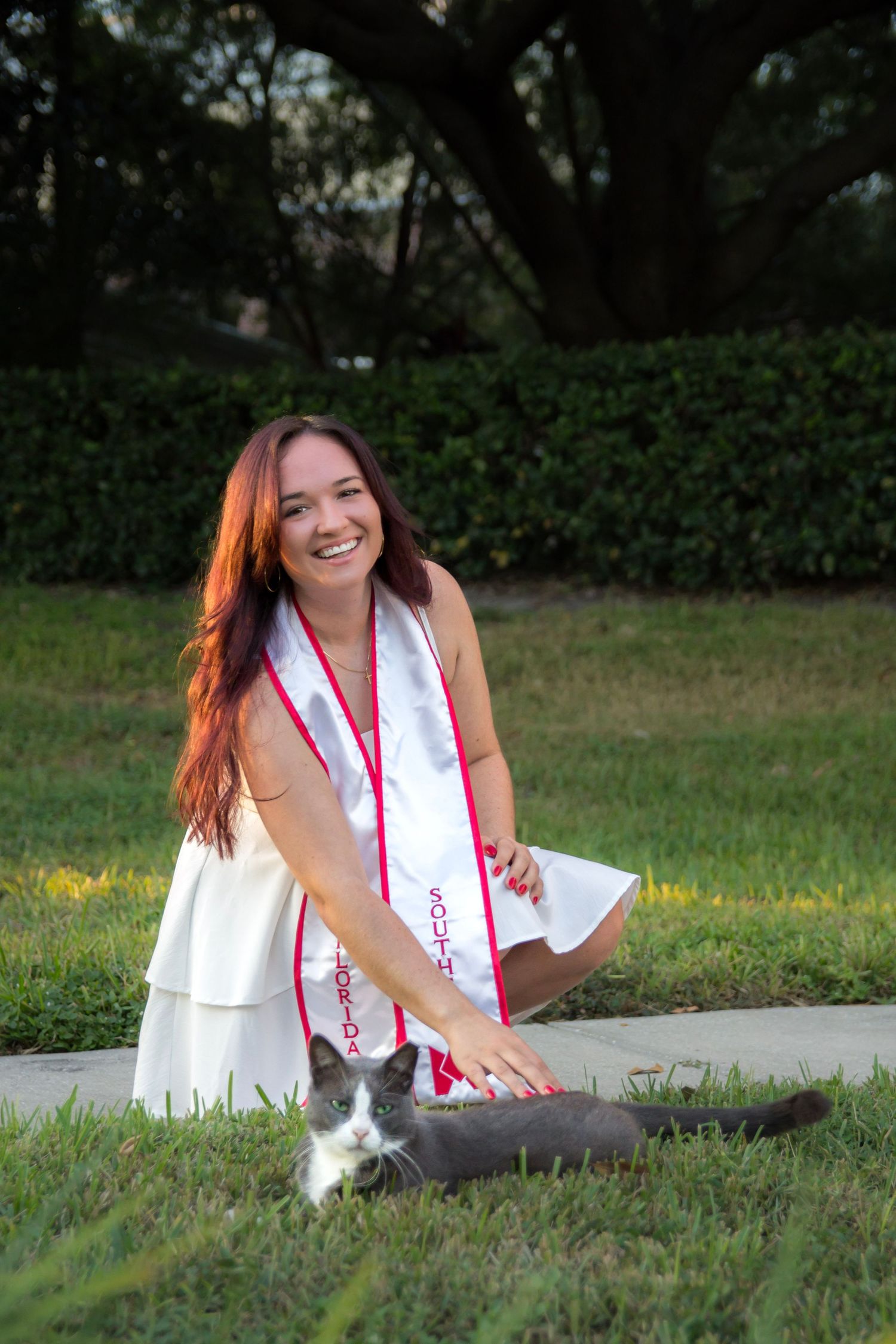 A graduate in white dress and red sash sits on grass during an outdoor photo session at sunset.