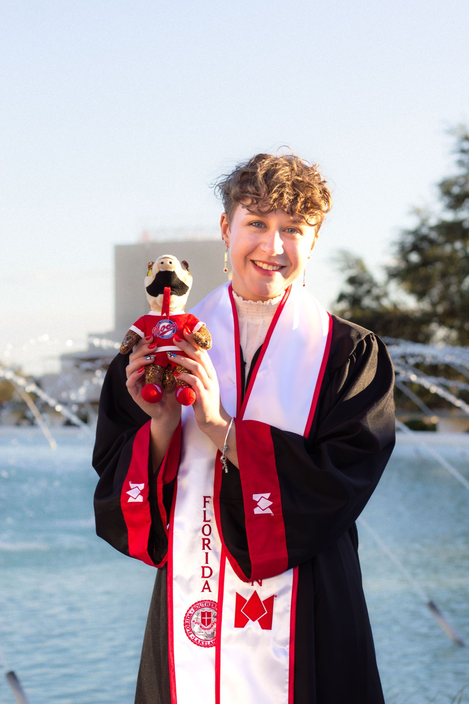 Graduate in black gown and red stole poses for photos by waterfront during graduation celebration.