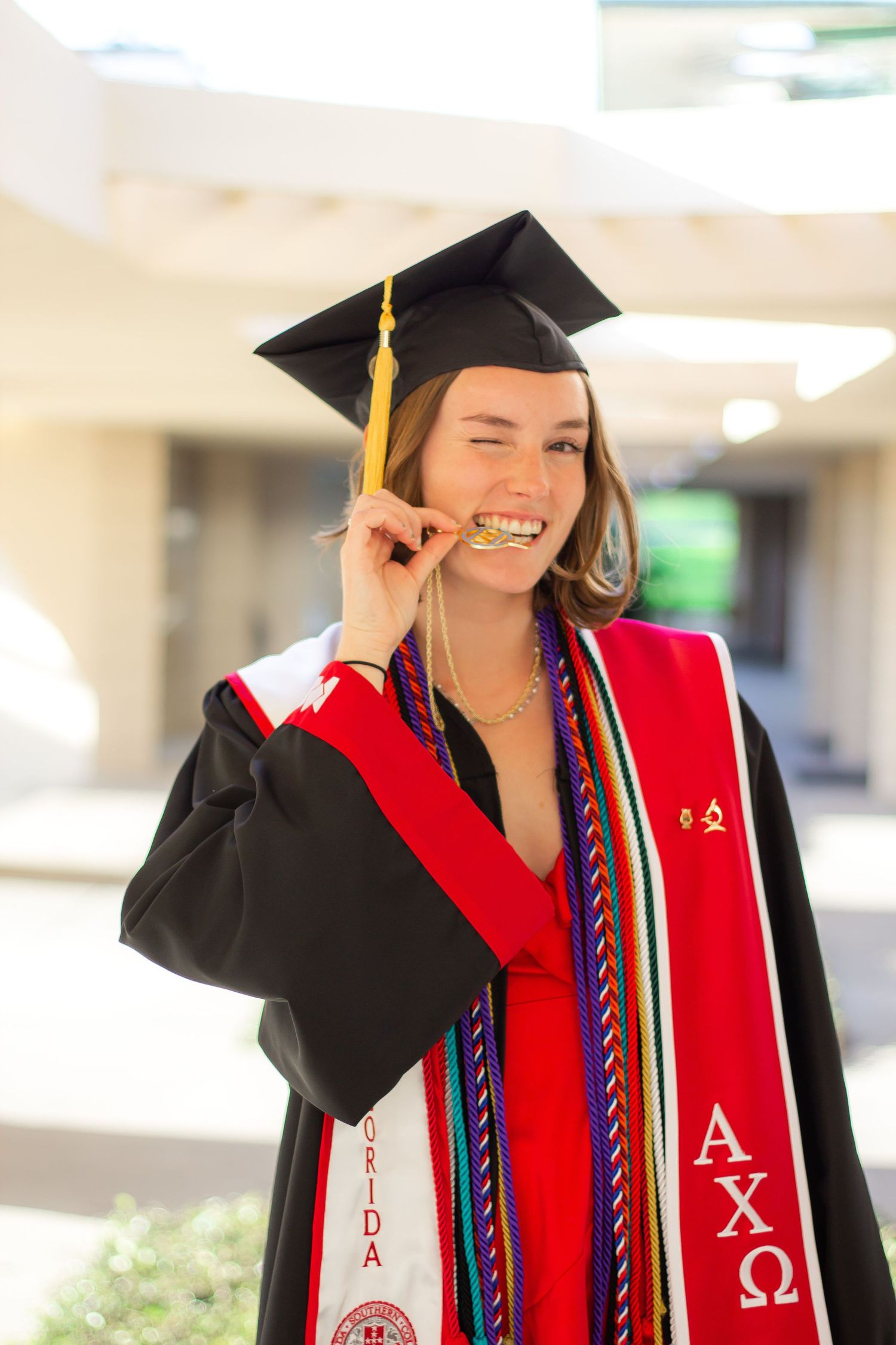 Graduate in black cap and gown with red stole celebrating at commencement ceremony outdoors.