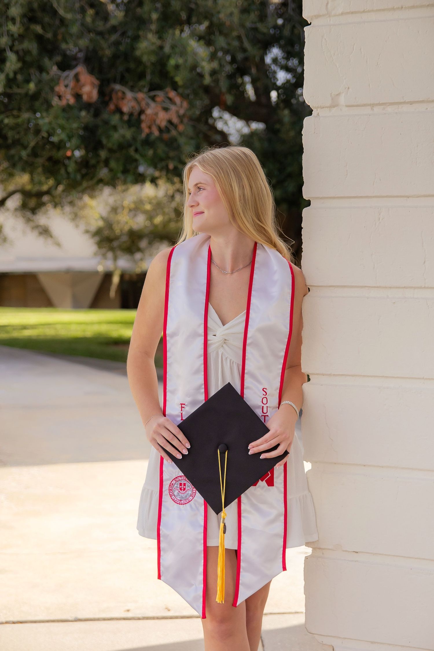 Graduate in pink and white stole poses with diploma against white column outdoors during graduation ceremony.