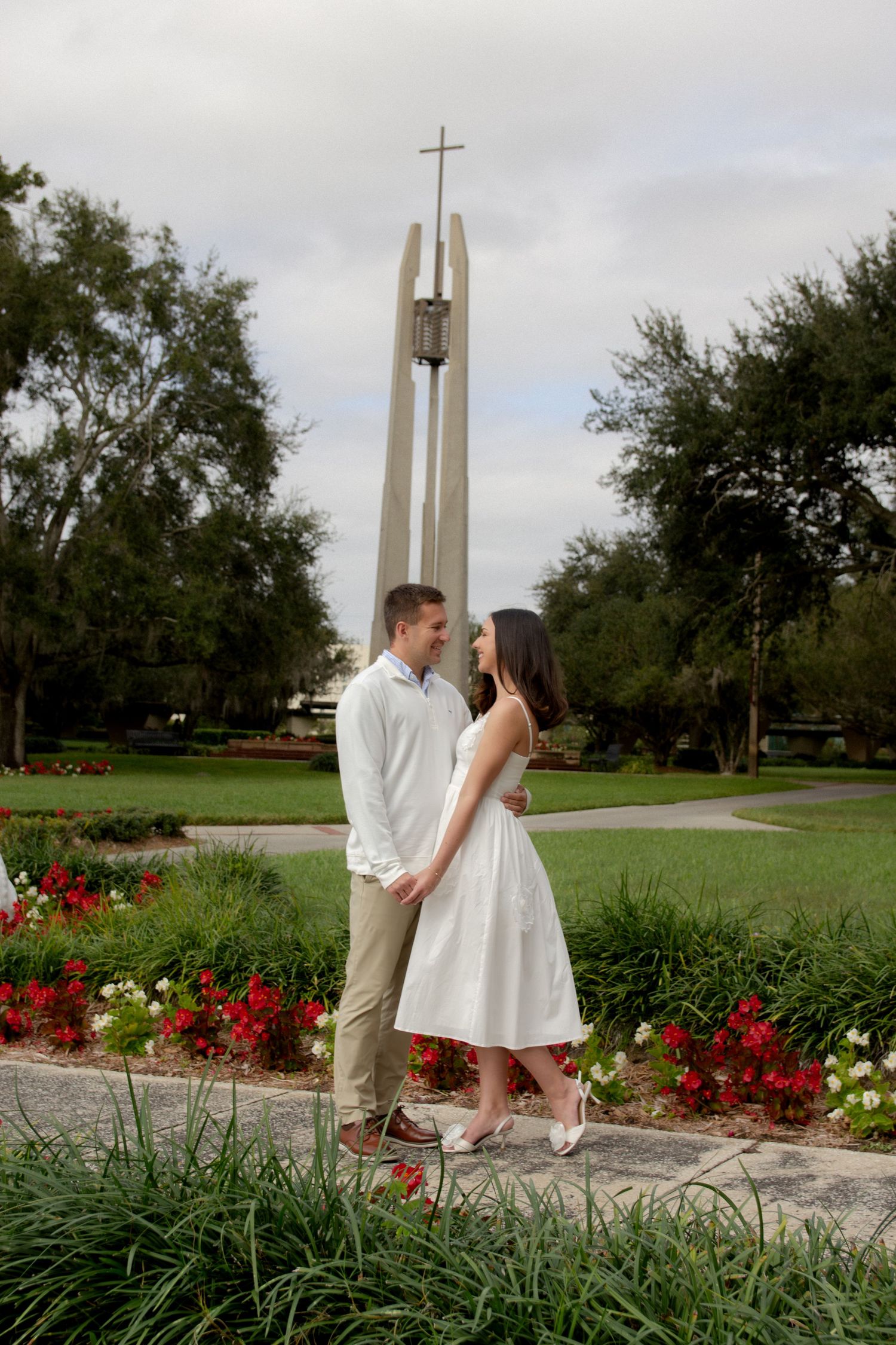 A couple sits on stone steps surrounded by red flowers and greenery near a modern church tower in a garden setting.