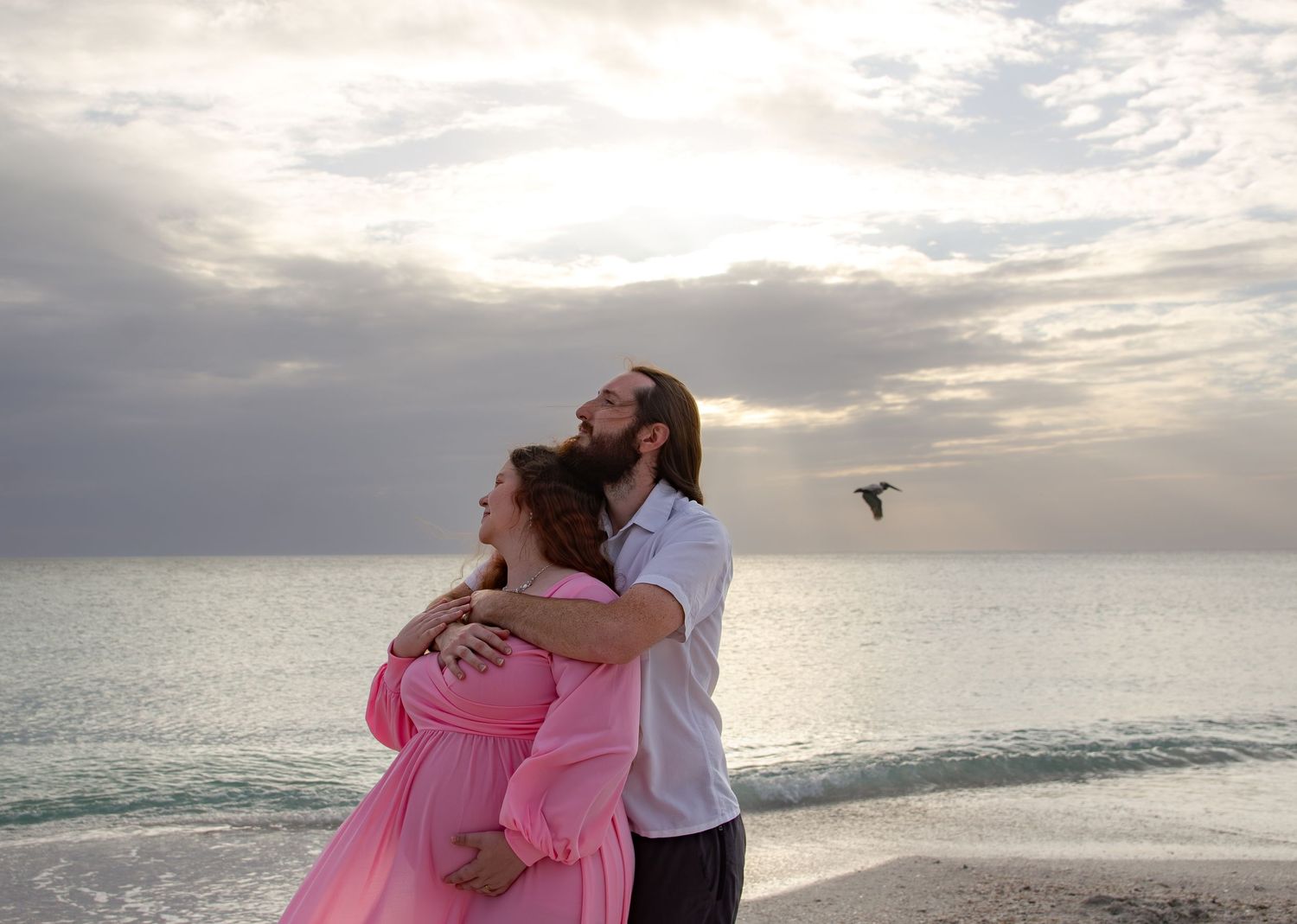 Couple embracing on beach at sunset, woman in pink maternity dress, man in white shirt, ocean backdrop.