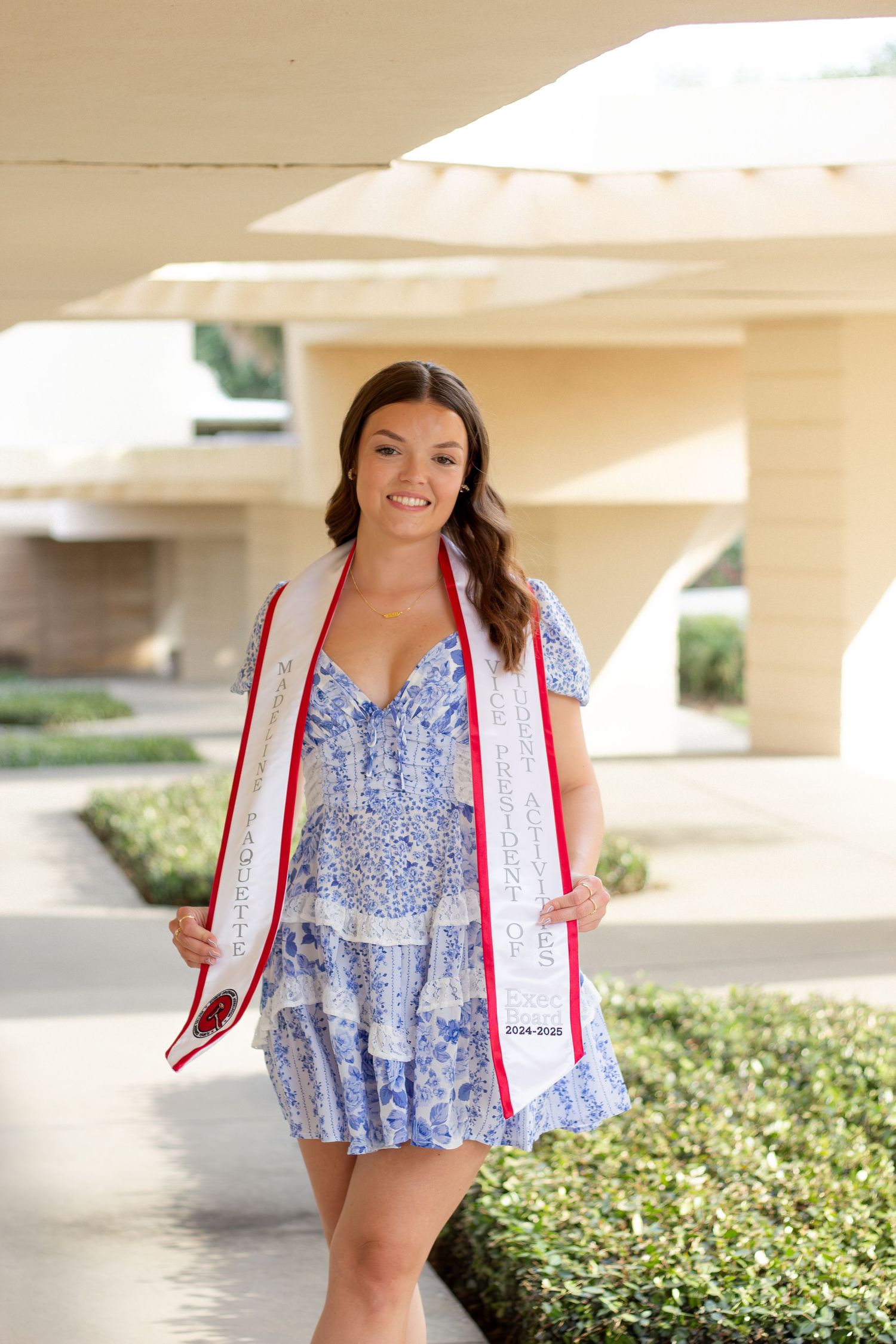 A graduate poses in floral dress and graduation stole in front of landscaped university grounds.