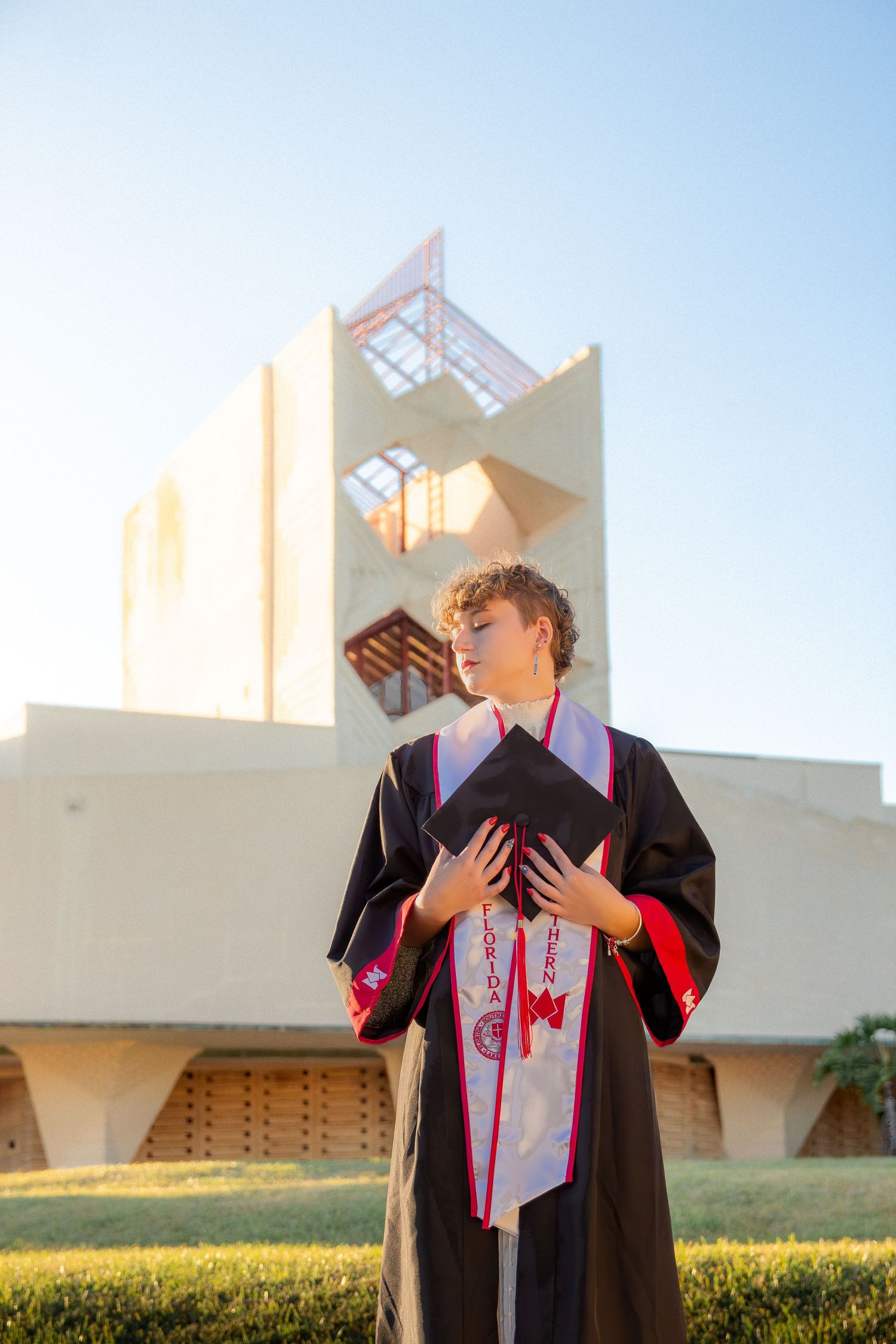 A graduate in cap and gown poses outdoors against a modern architectural backdrop during a sunny day.
