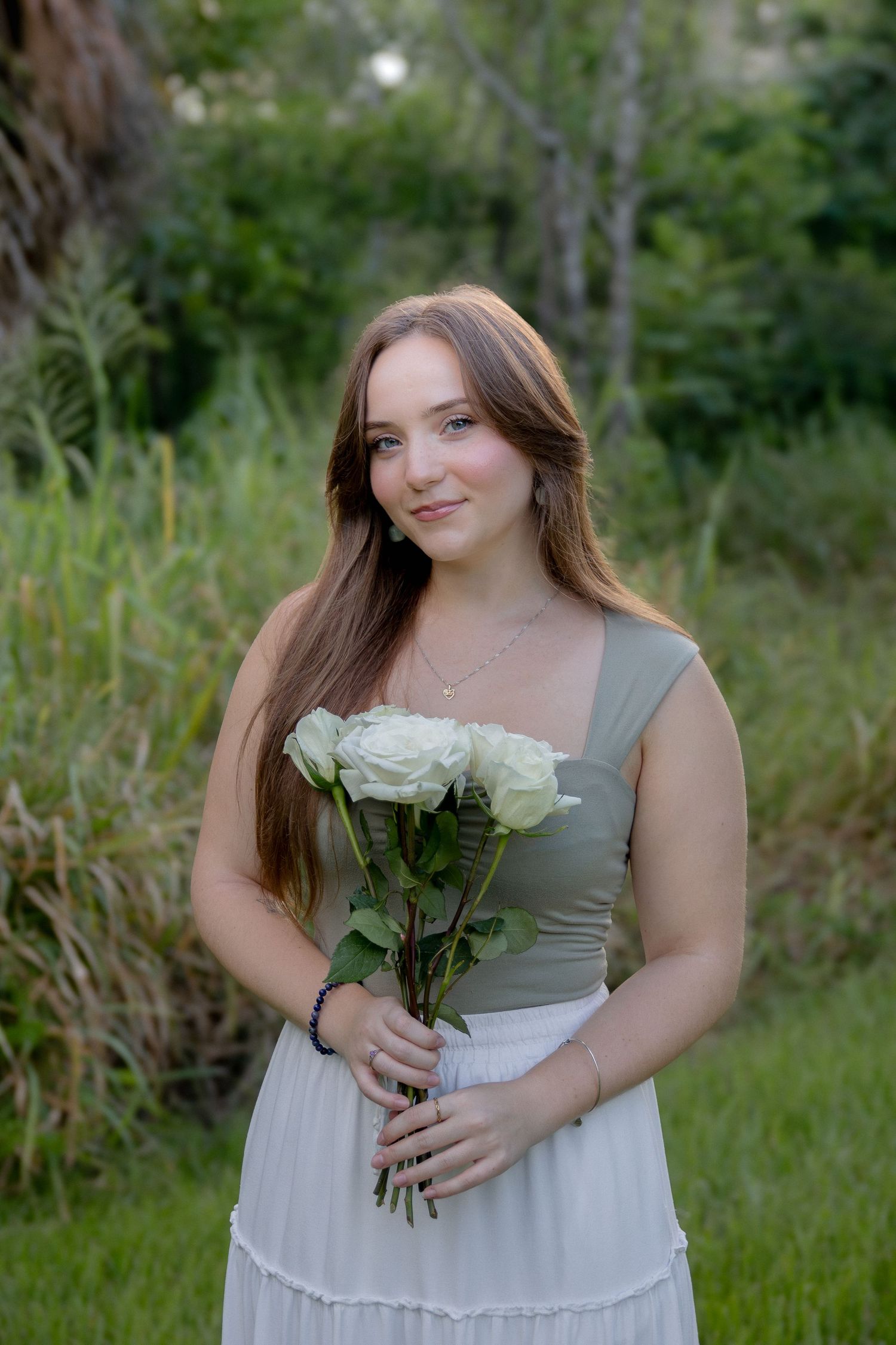 A woman in a white and gray dress holds white roses while standing in a grassy outdoor setting.