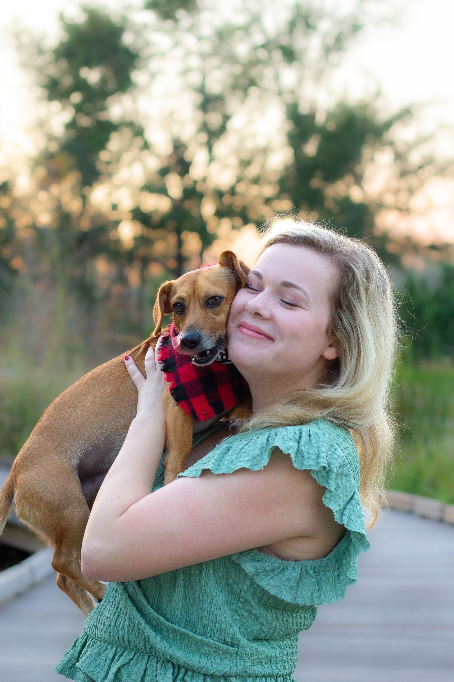 A person in a mint green top cuddles and plays with a small brown dog outdoors at sunset.
