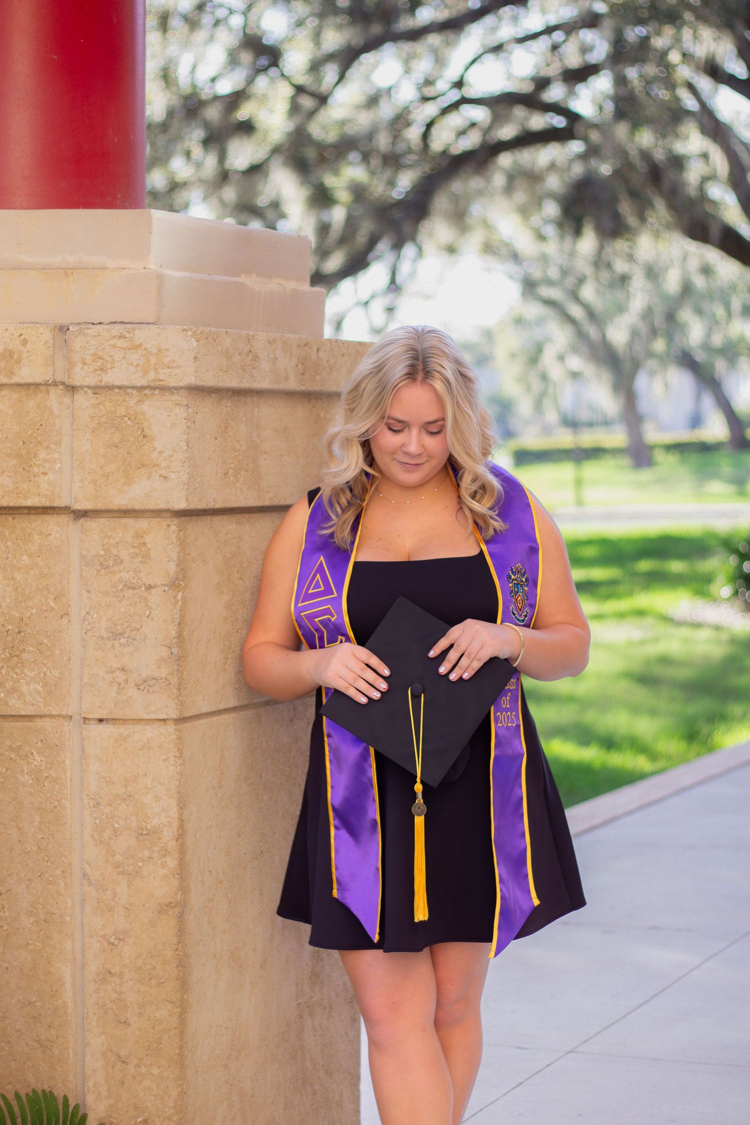 Graduate in black dress and purple stole looks down at their diploma during outdoor graduation photo session.