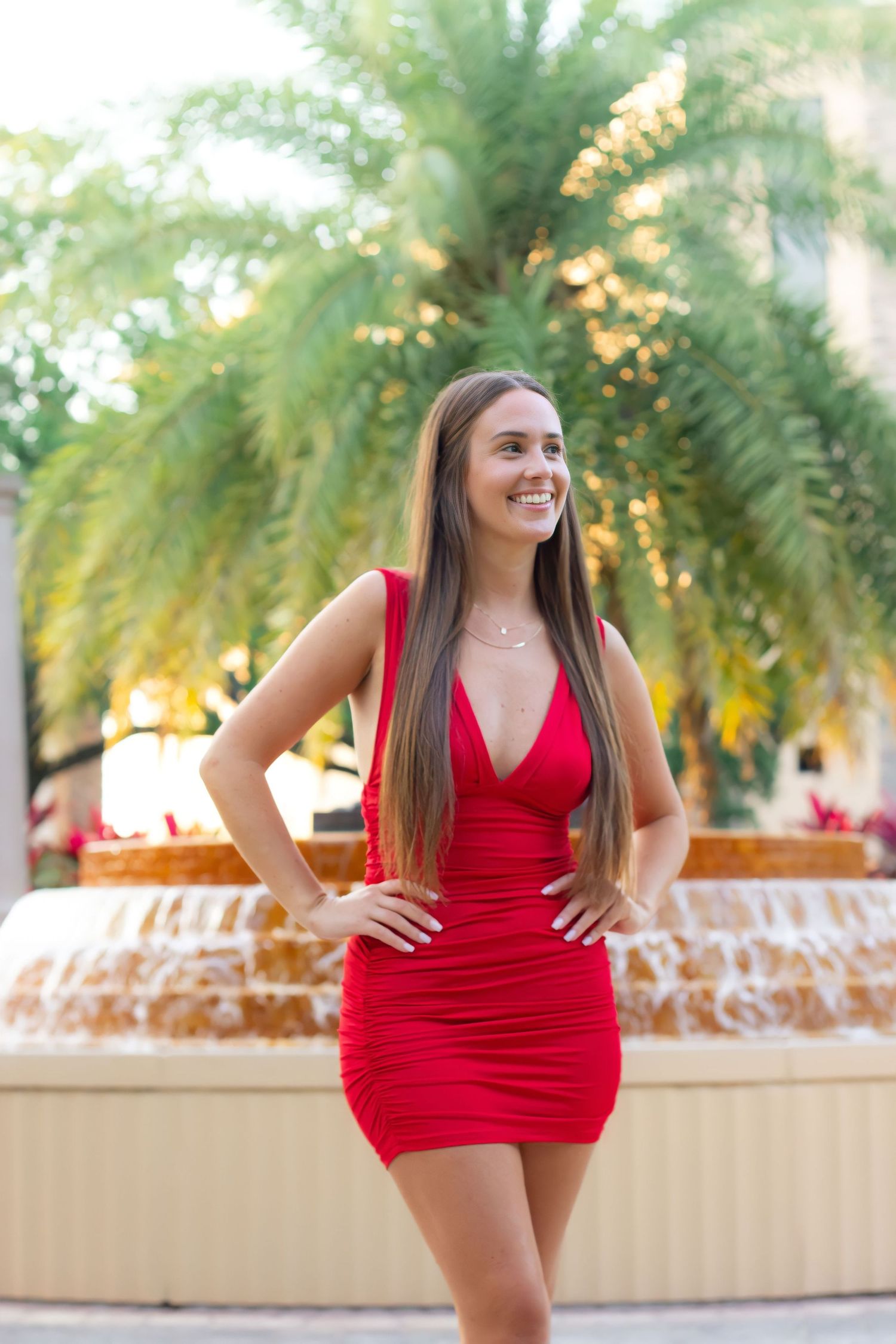 A woman in a red dress poses in front of palm trees and a fountain at a resort.