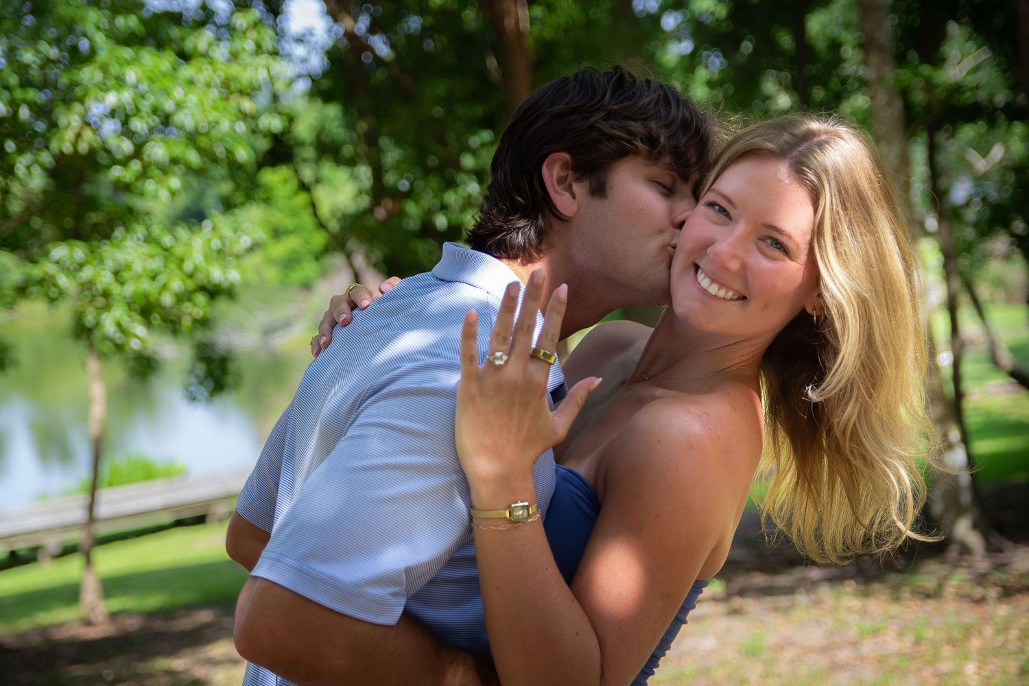 Happy couple in park, woman showing engagement ring while man kisses her cheek, trees and lake in background.