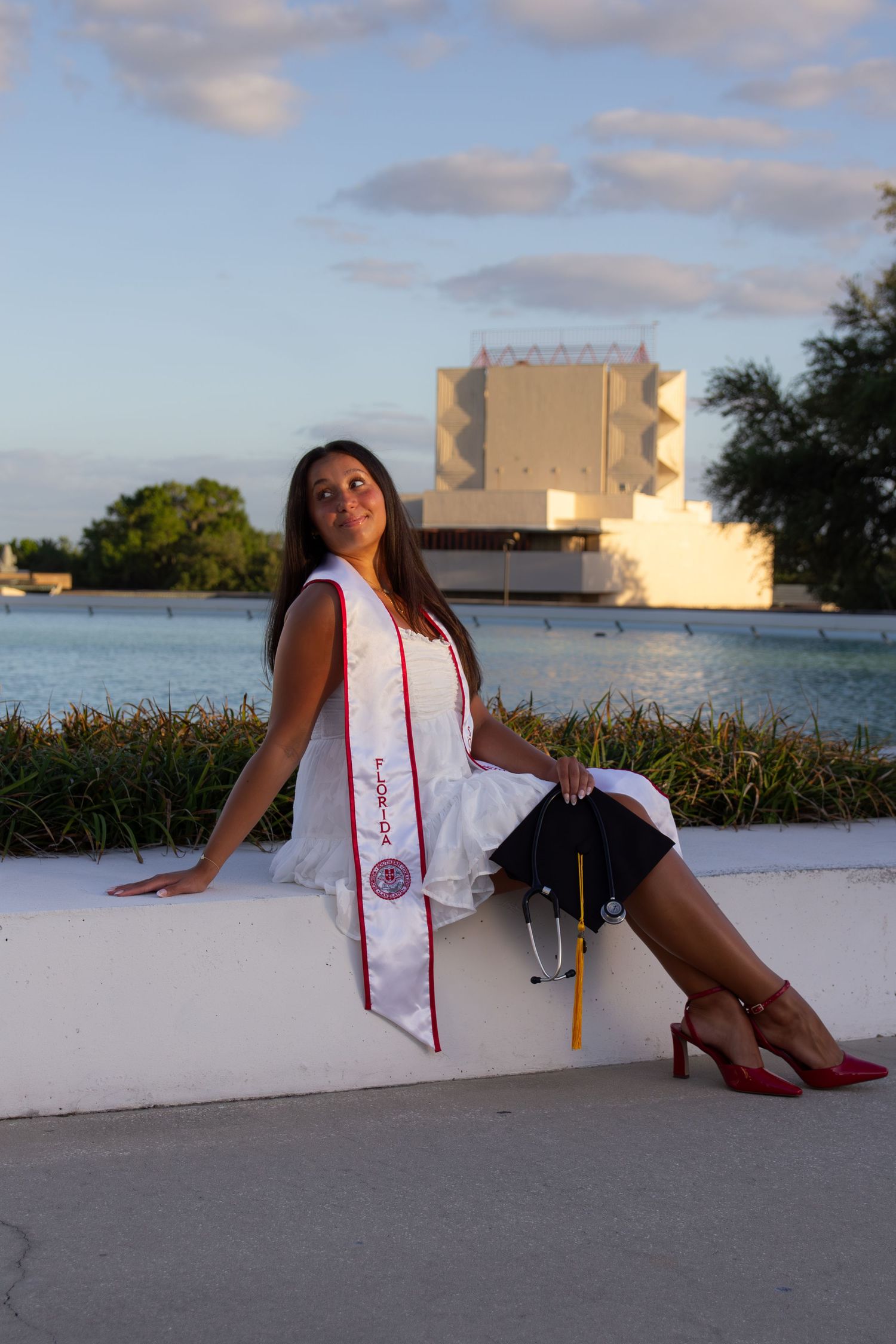 Young woman in white graduation gown and red sash sits outdoors near a lake, holding cap, wearing red heels.