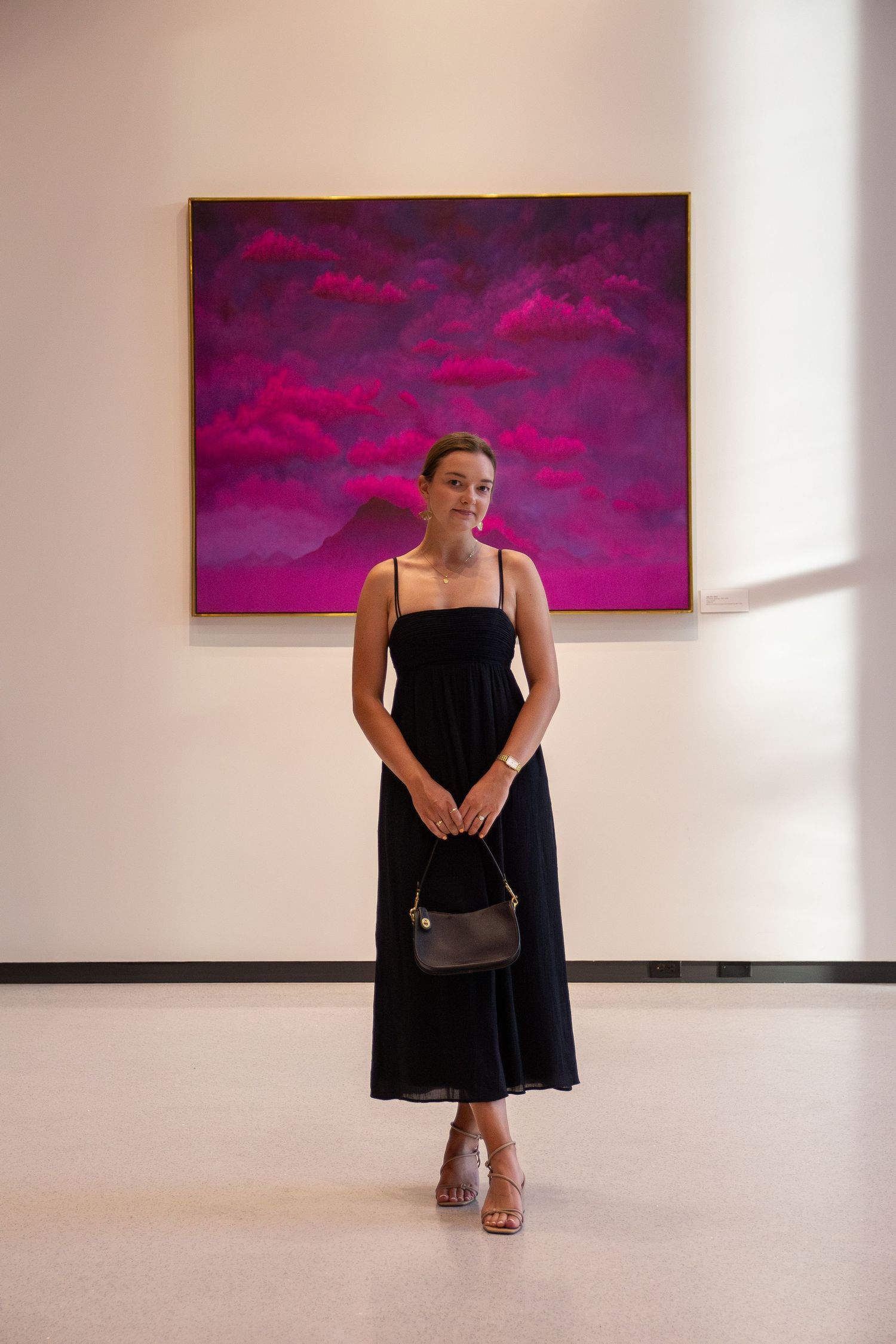 Person in black dress standing in art gallery with vibrant pink paintings on white walls.