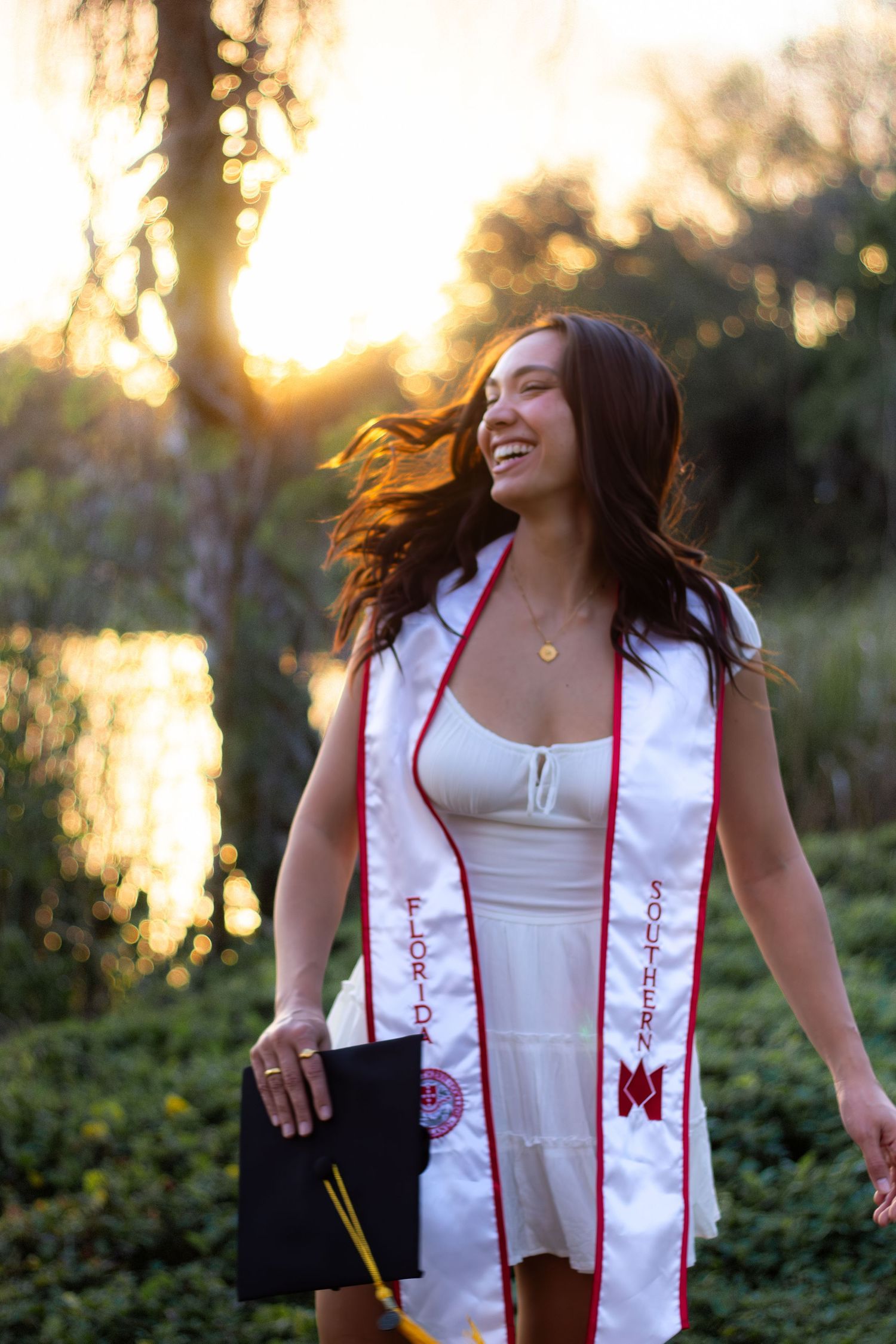 Joyful graduation portraits at sunset wearing white stole with red accents.
