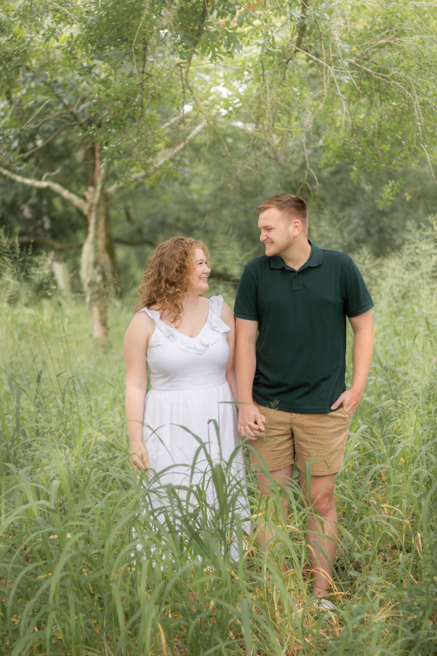 A couple in white and dark casual outfits walk through a lush green meadow filled with tall grass and trees.