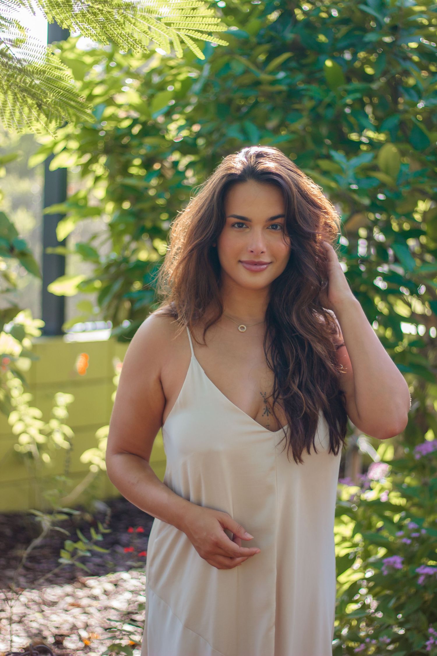 Woman in white sleeveless top poses outdoors against lush green foliage background in summer setting.