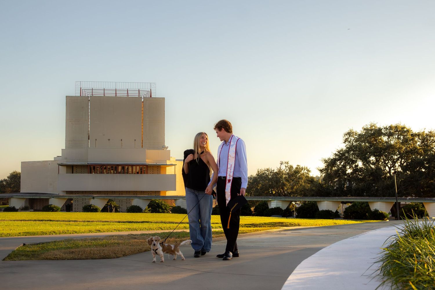 People walk along a scenic pathway at sunset with golden fields and buildings visible in the distance.