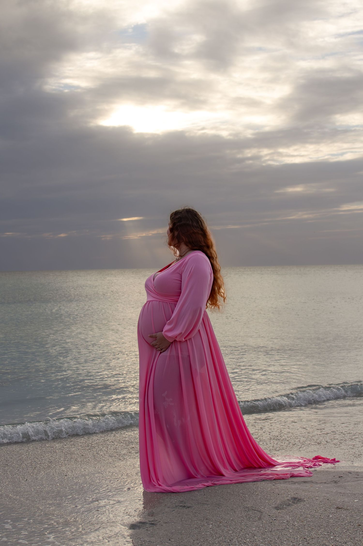 Pregnant woman in pink maternity gown standing on beach at sunset, cradling baby bump with ocean waves behind her.