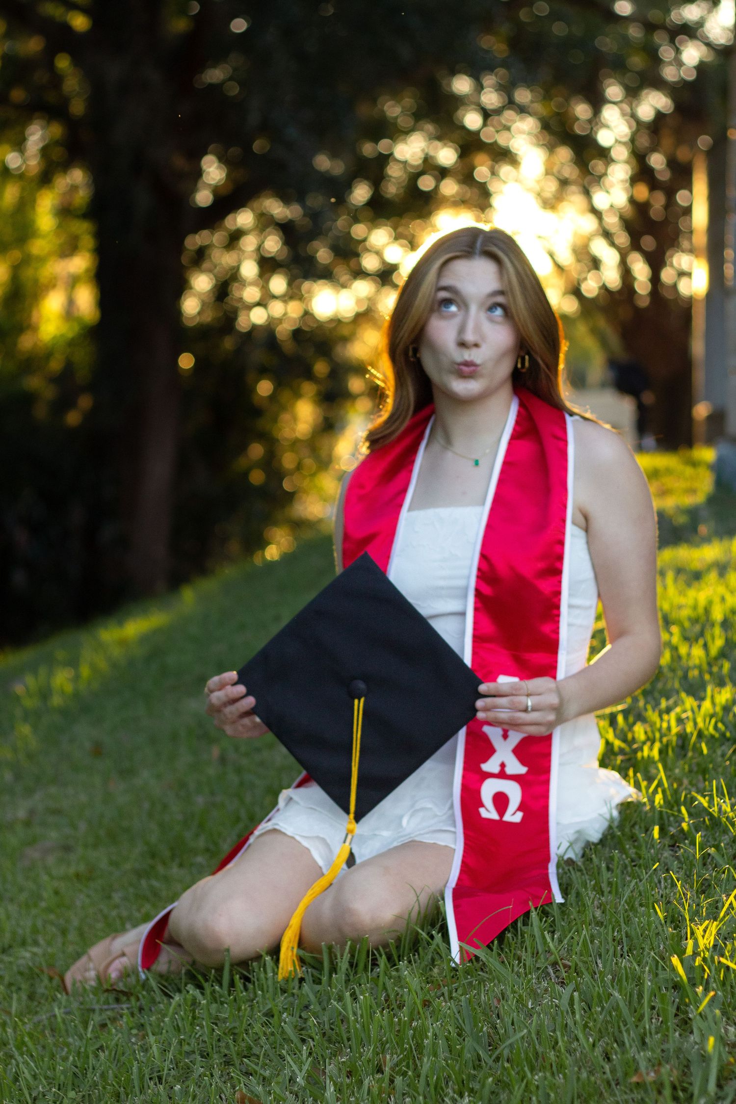 Graduate in white dress with red stole and black cap poses on grass at sunset during outdoor graduation photoshoot.