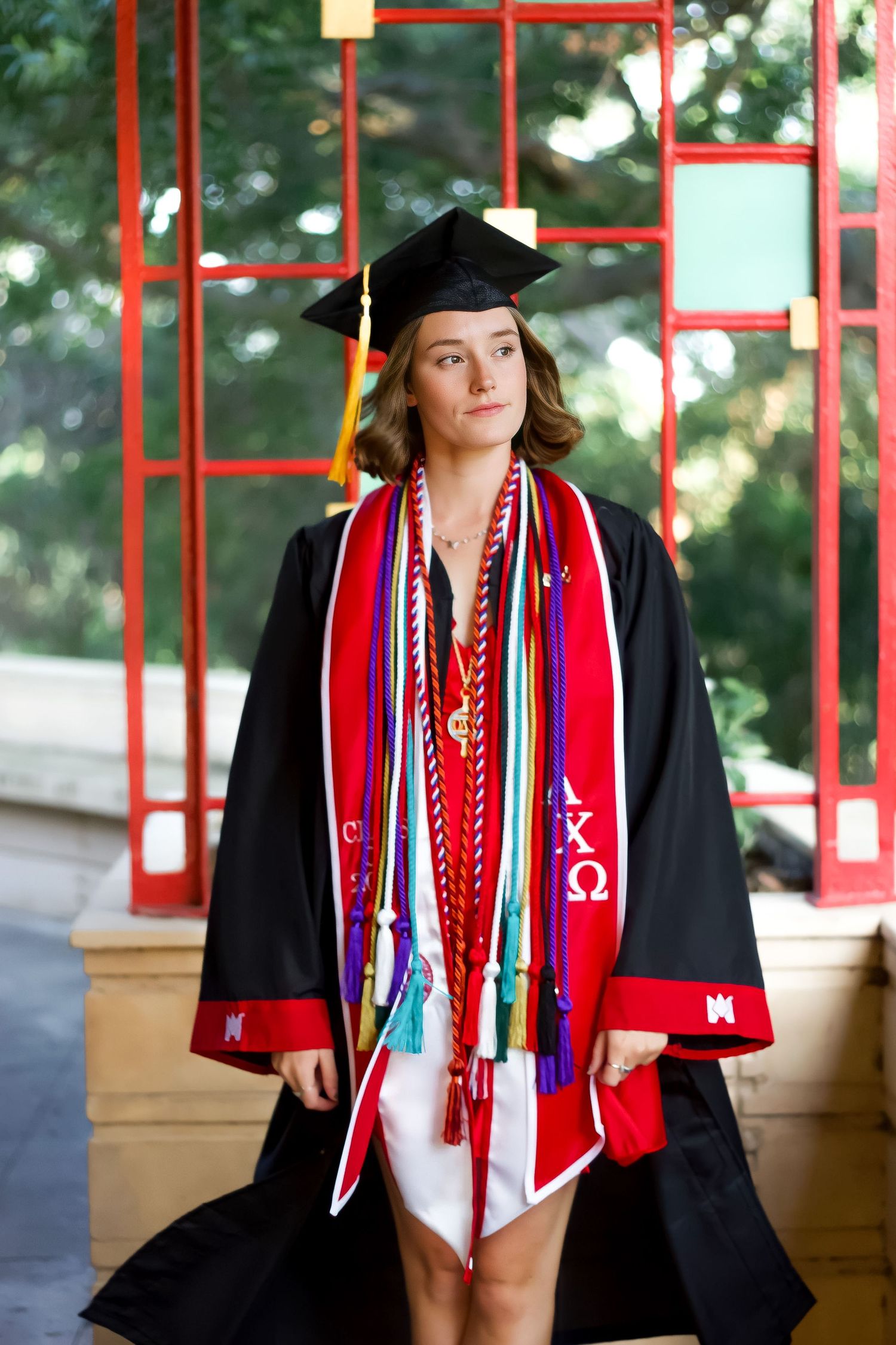 Graduate wearing black cap and gown with red stoles and colorful graduation cords during commencement ceremony.