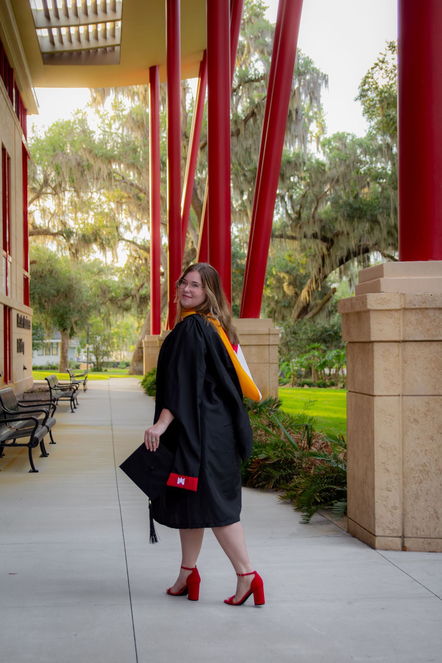 Young woman in black graduation gown and red heels poses on campus walkway with red columns and trees.