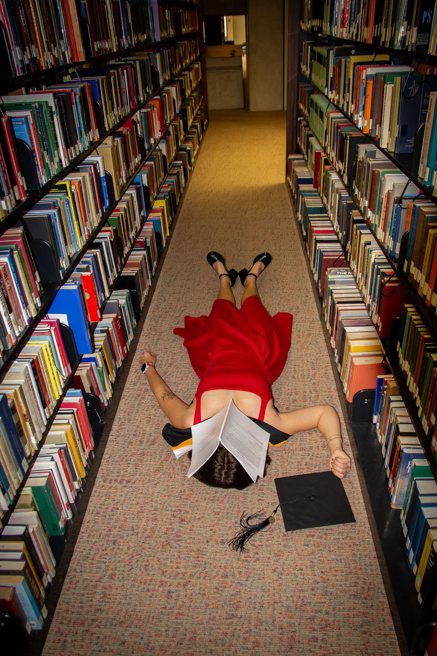 A graduate in red dress lies on library floor between bookshelves, holding diploma with mortarboard cap nearby.