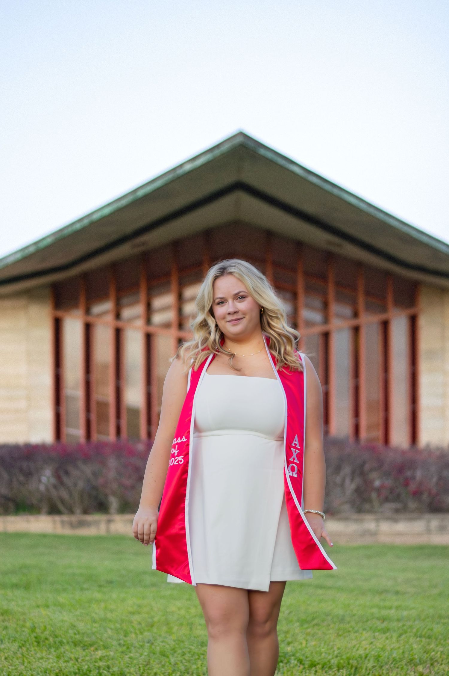 A person in a red blazer and white dress poses outdoors in front of a modern architectural building.