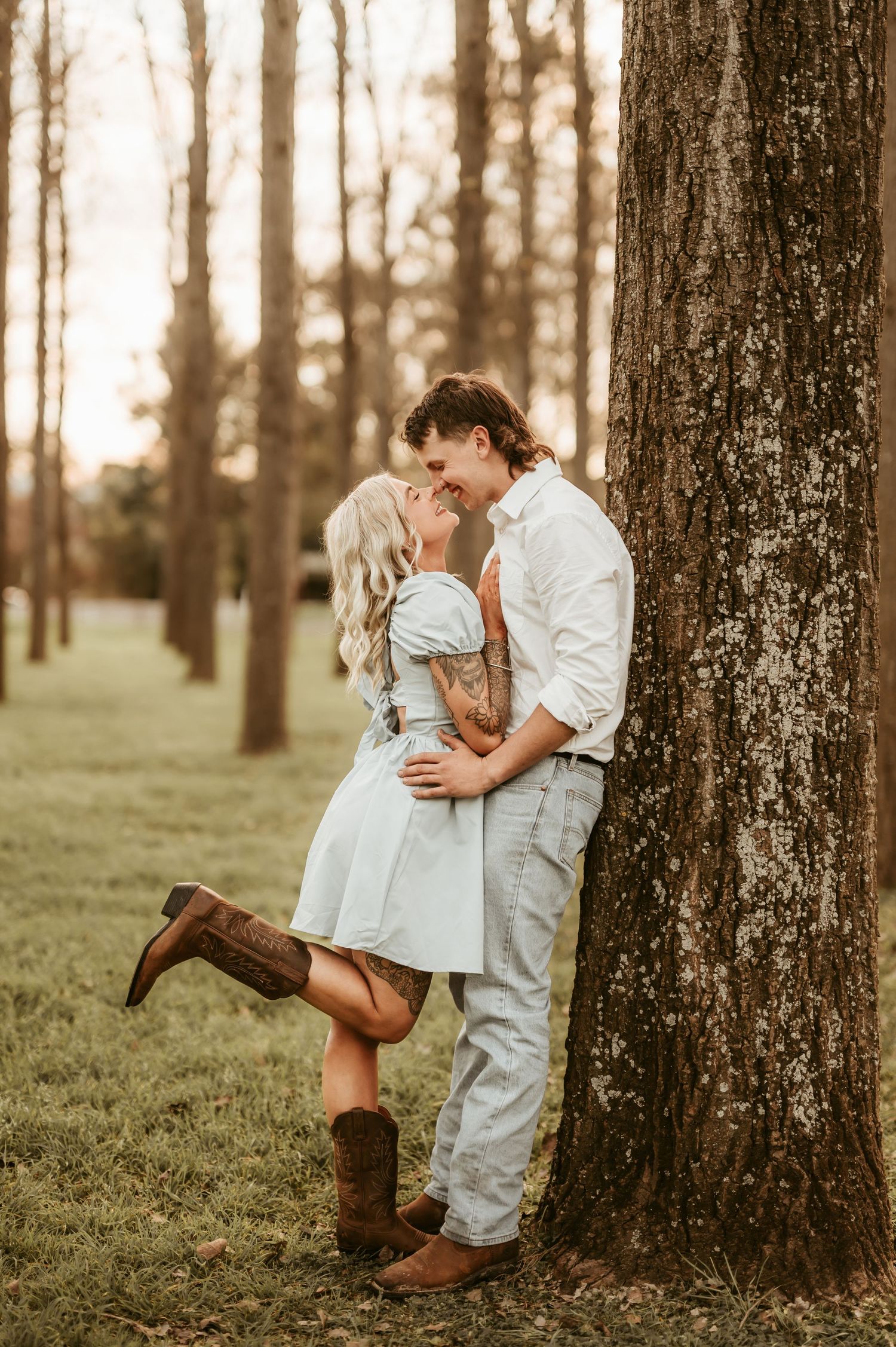 Romantic couple in white clothing and boots poses against tree trunks in a wooded setting.