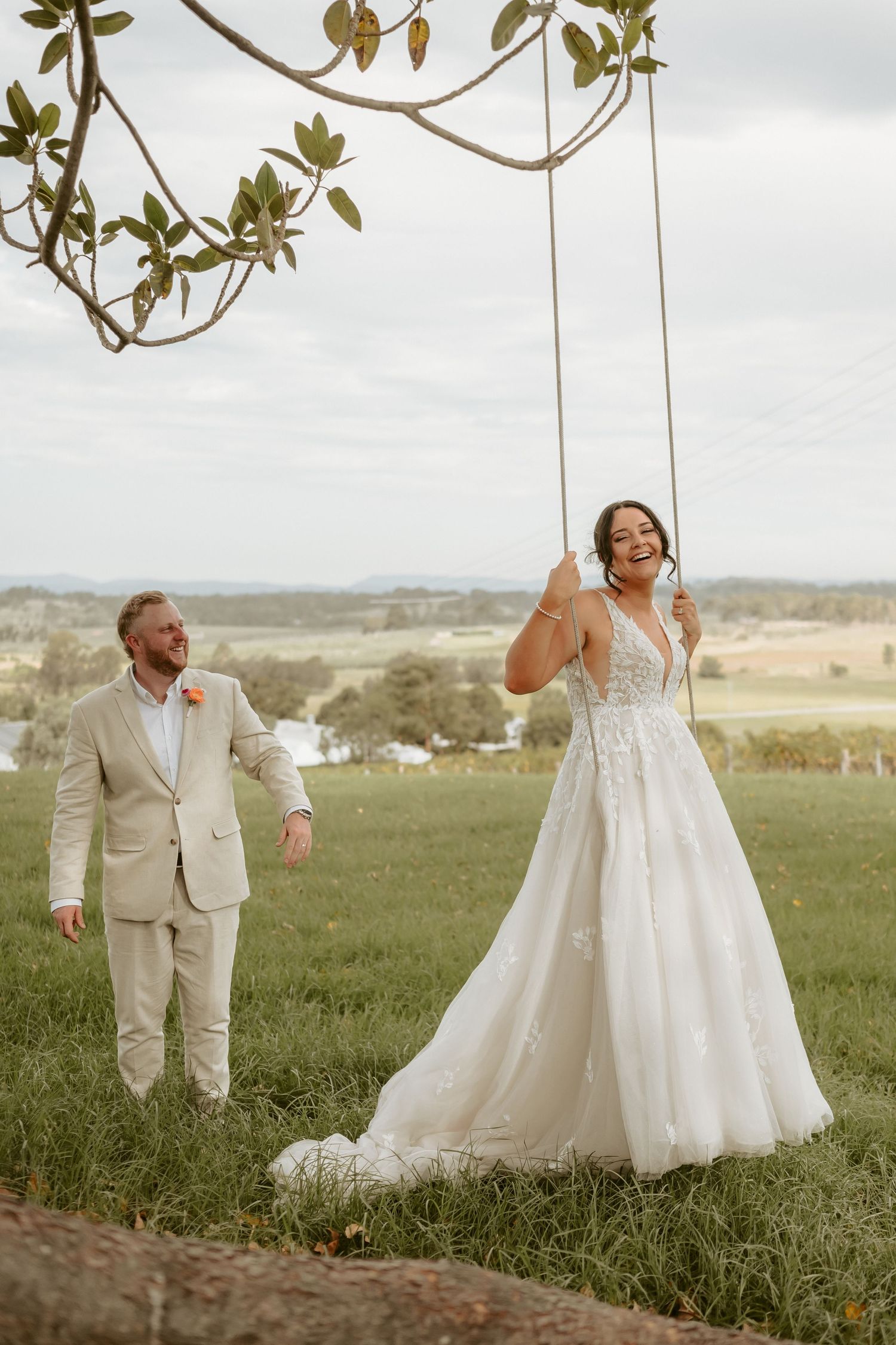 Couple in wedding attire playfully posing in a green field with mountains in background.