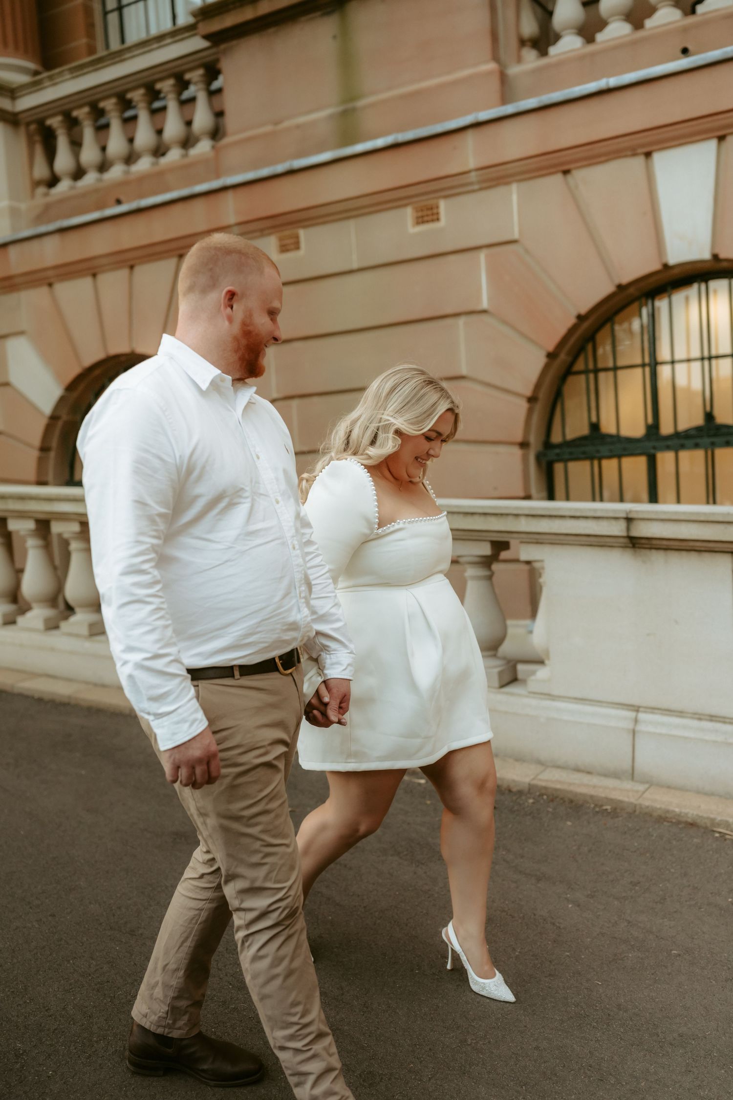 A couple in white and beige attire walking together along a classic stone building exterior, showcasing elegant style.
