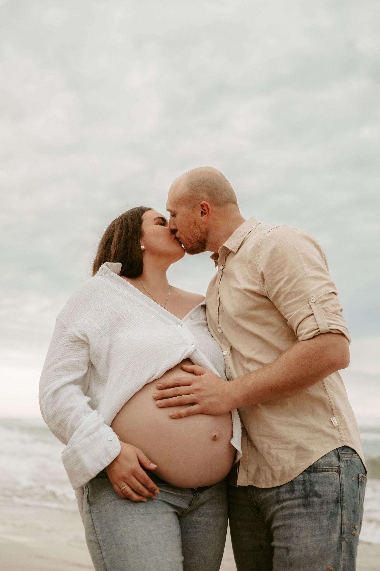 An intimate maternity photo of a couple sharing a tender moment on a cloudy beach.