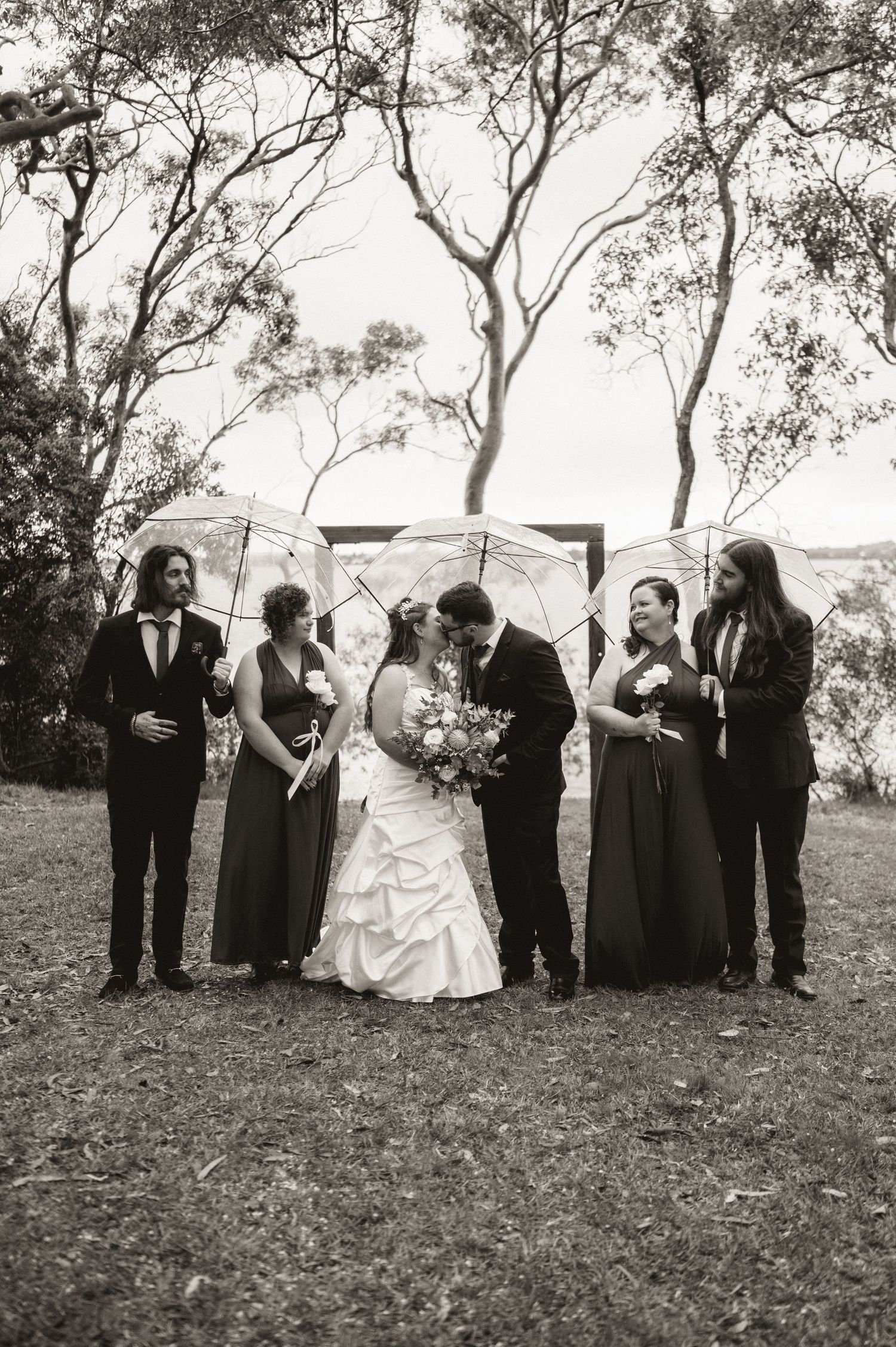 Wedding party poses together on a grassy hillside with trees in the background.