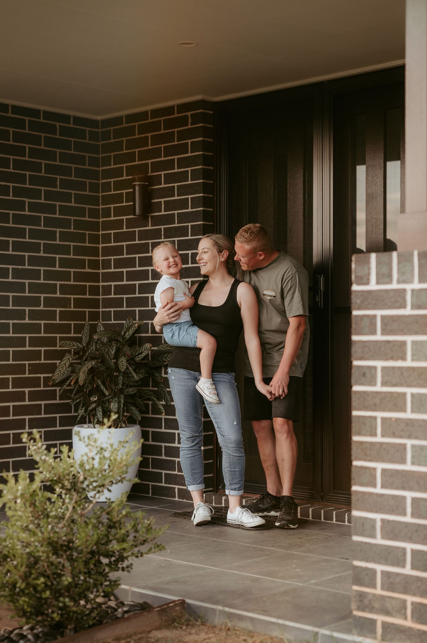 Family members share a playful moment on a brick porch with potted plants.