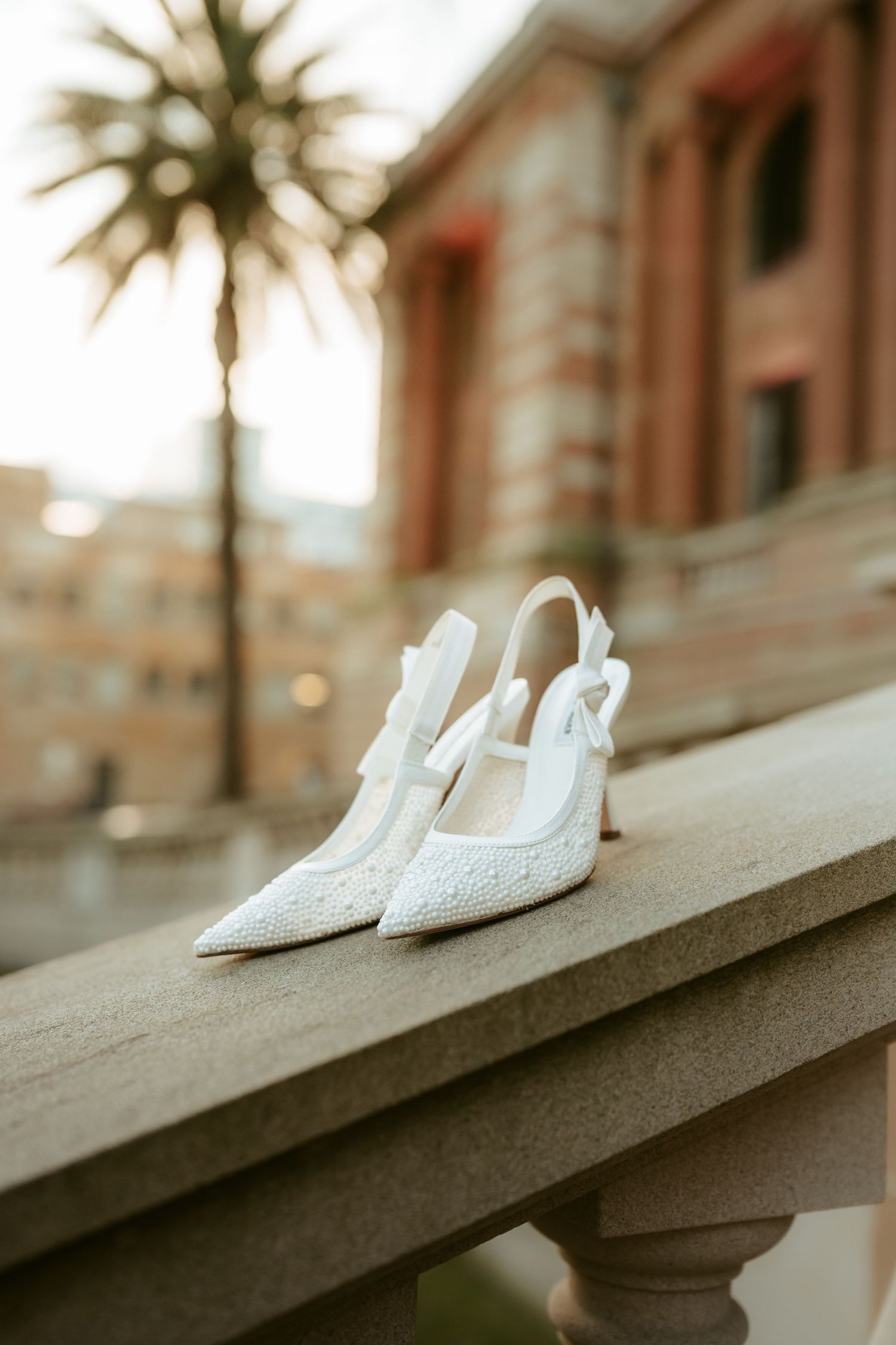 White bridal shoes sit elegantly on a concrete ledge with palm trees and architecture visible in the soft background.