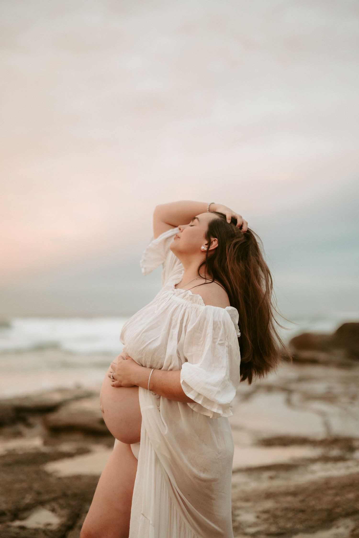 A dramatic maternity photo of a woman in white dress posing on coastal rocks at sunset.