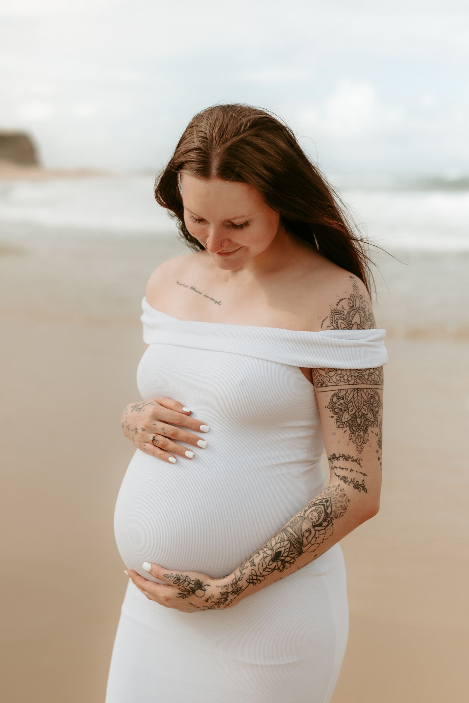 An expectant mother in a white dress shows her pregnant belly against a beach backdrop.
