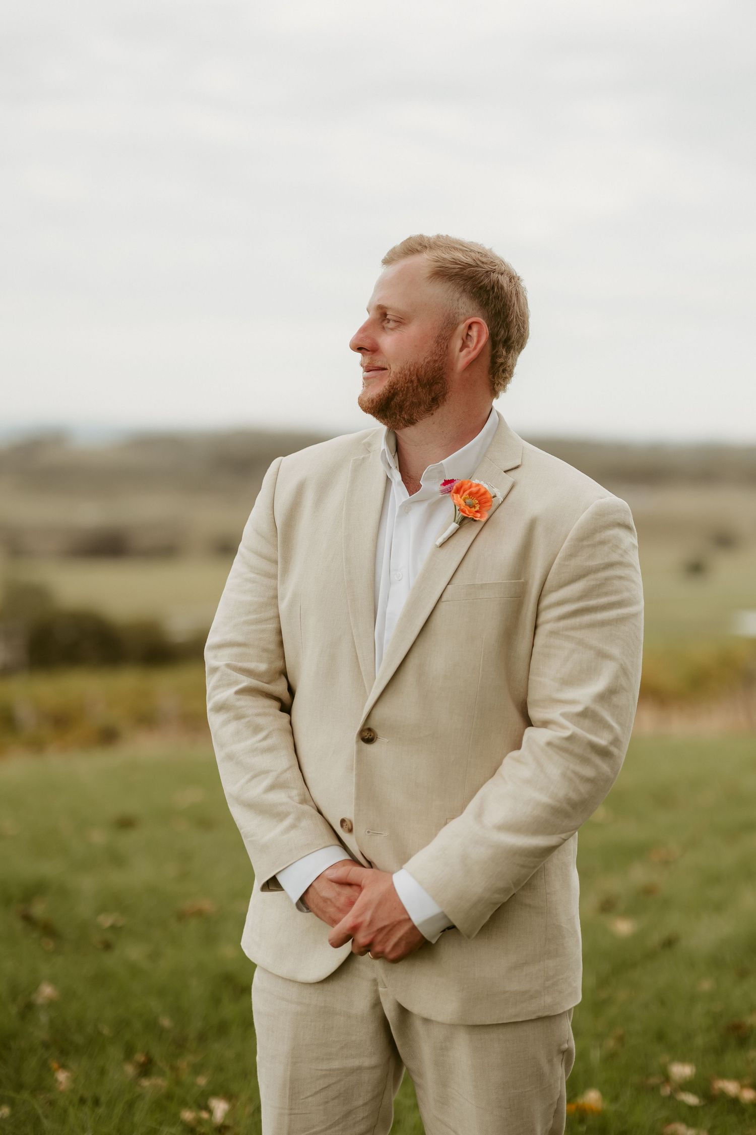 Groom in light beige suit with orange boutonniere poses in outdoor field setting.