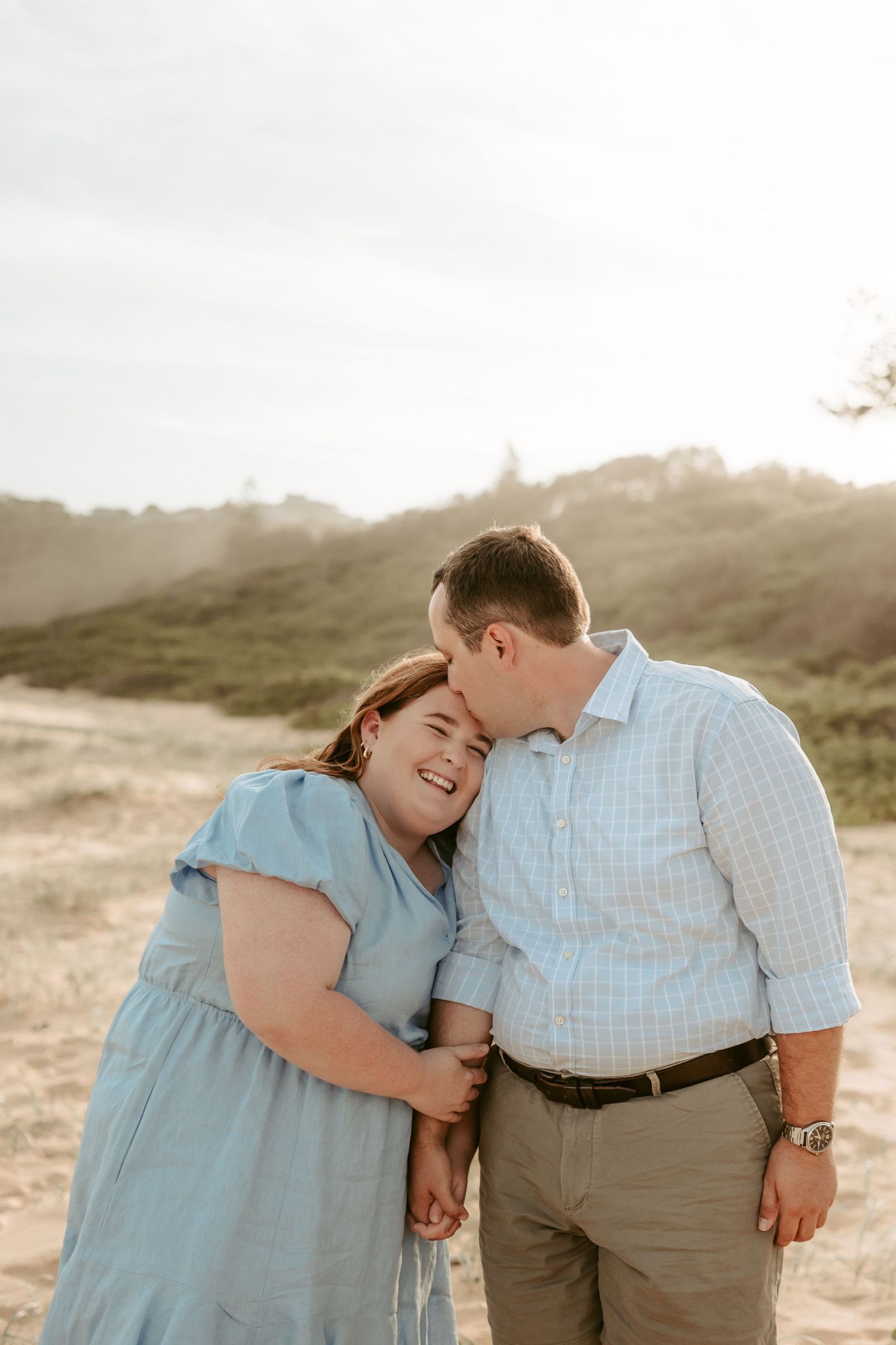 Loving couple in blue and white outfits embracing against a scenic hillside backdrop.