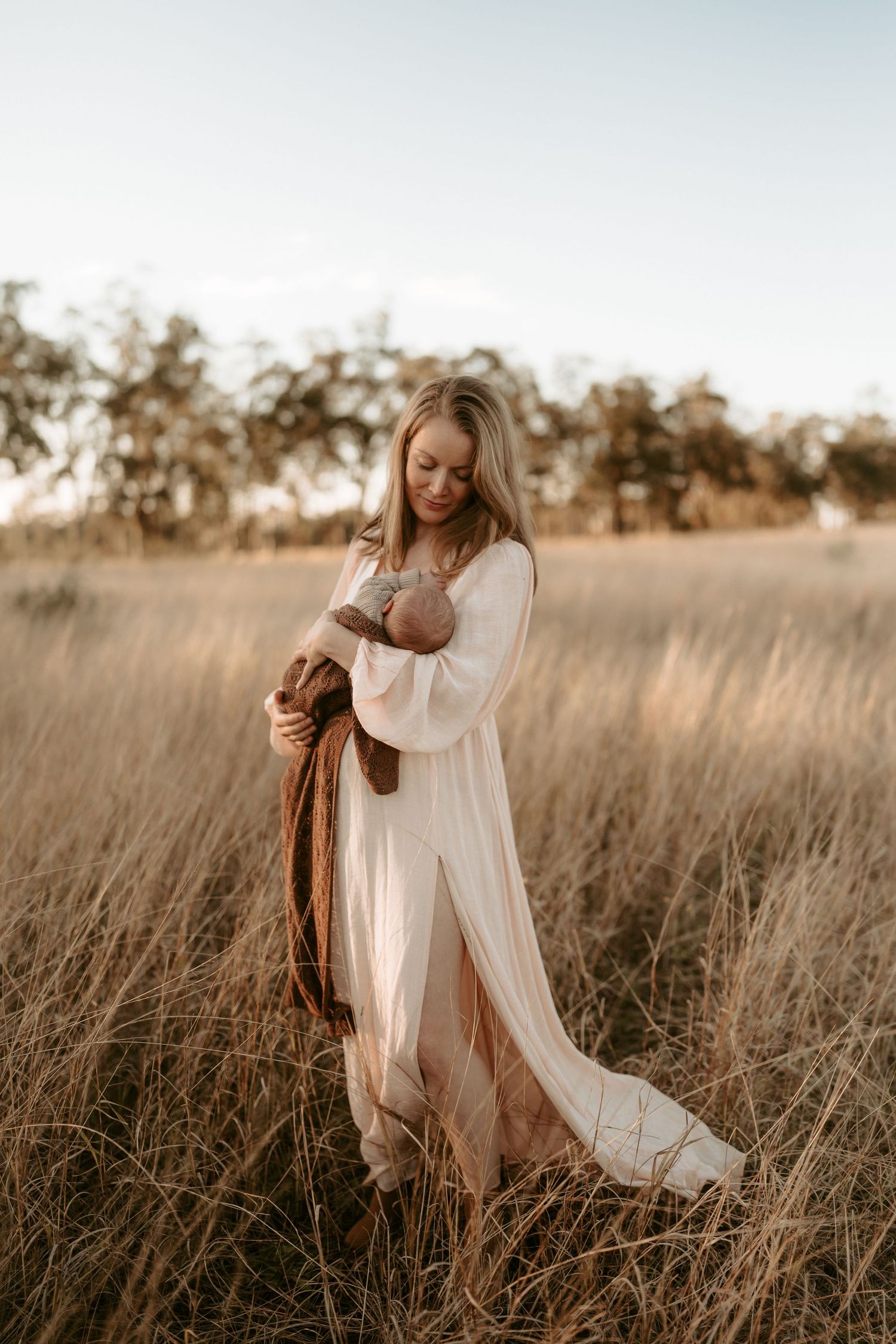 Mother in flowing white dress holding baby in rustic outdoor setting at golden hour.