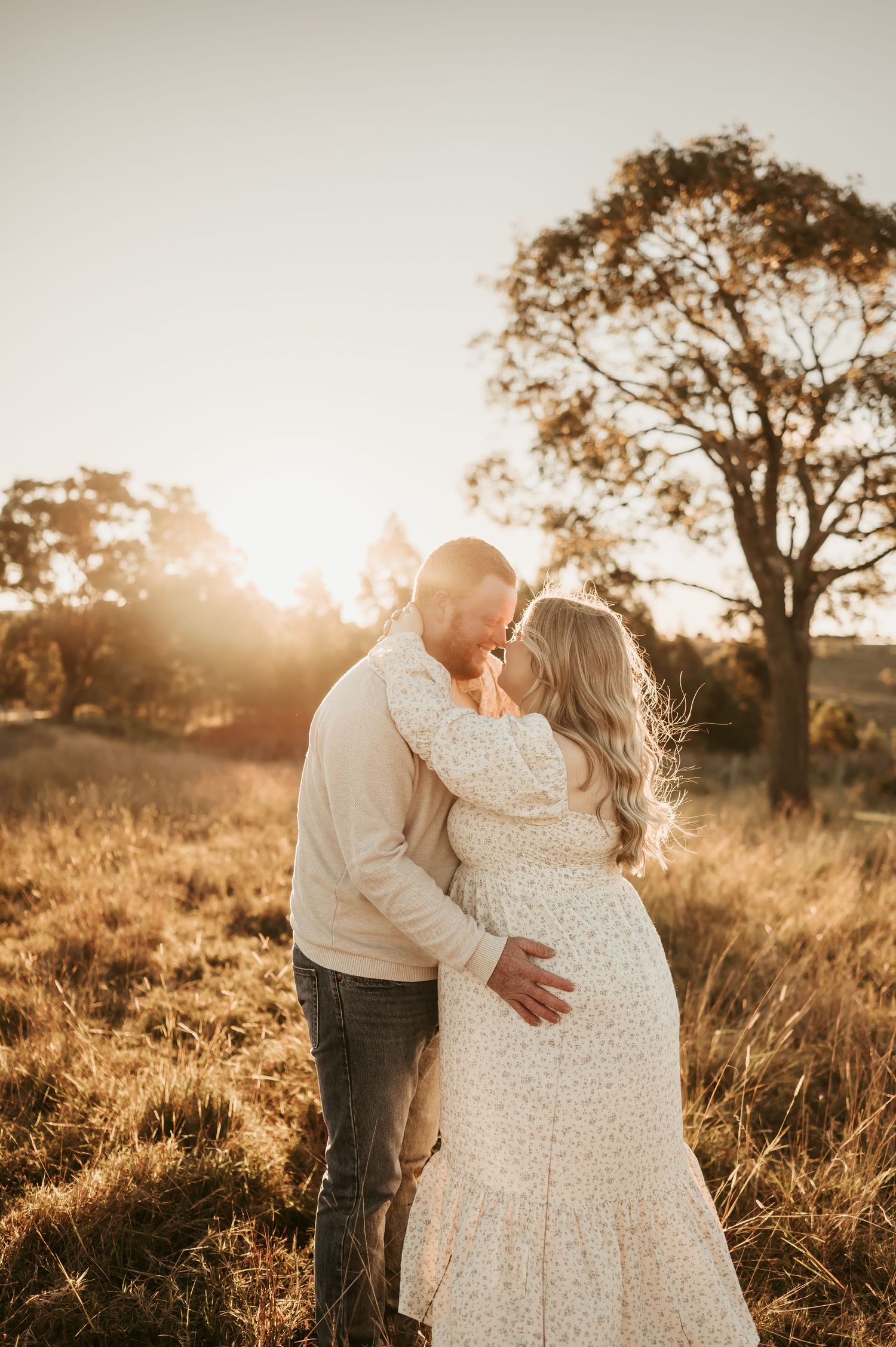 Couple sharing an intimate moment in a sunlit field with trees in the background.