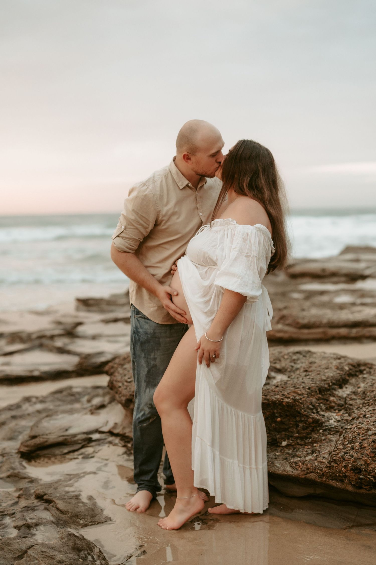 A romantic maternity photo of a couple embracing on rocky coastal shores at sunset.