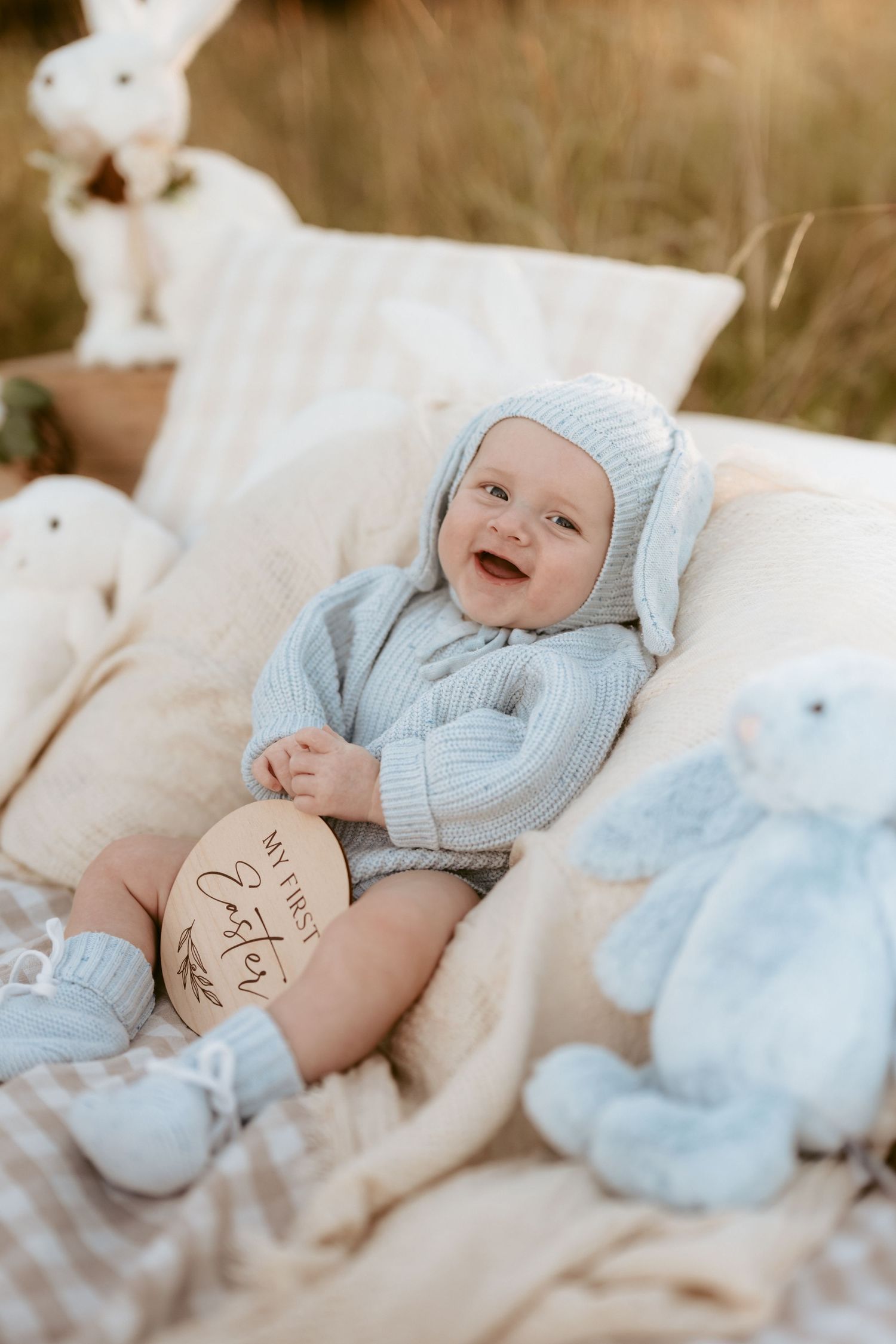 Baby wearing blue outfit smiles during Easter-themed photoshoot on white blanket.