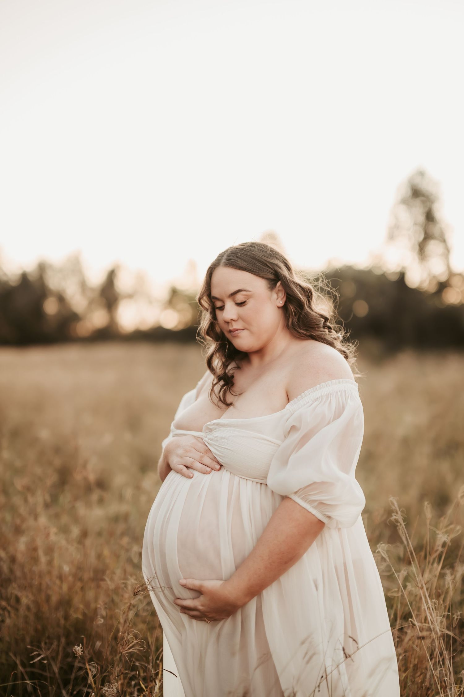 Expectant mother in white dress tenderly cradling her belly in a sunlit meadow.