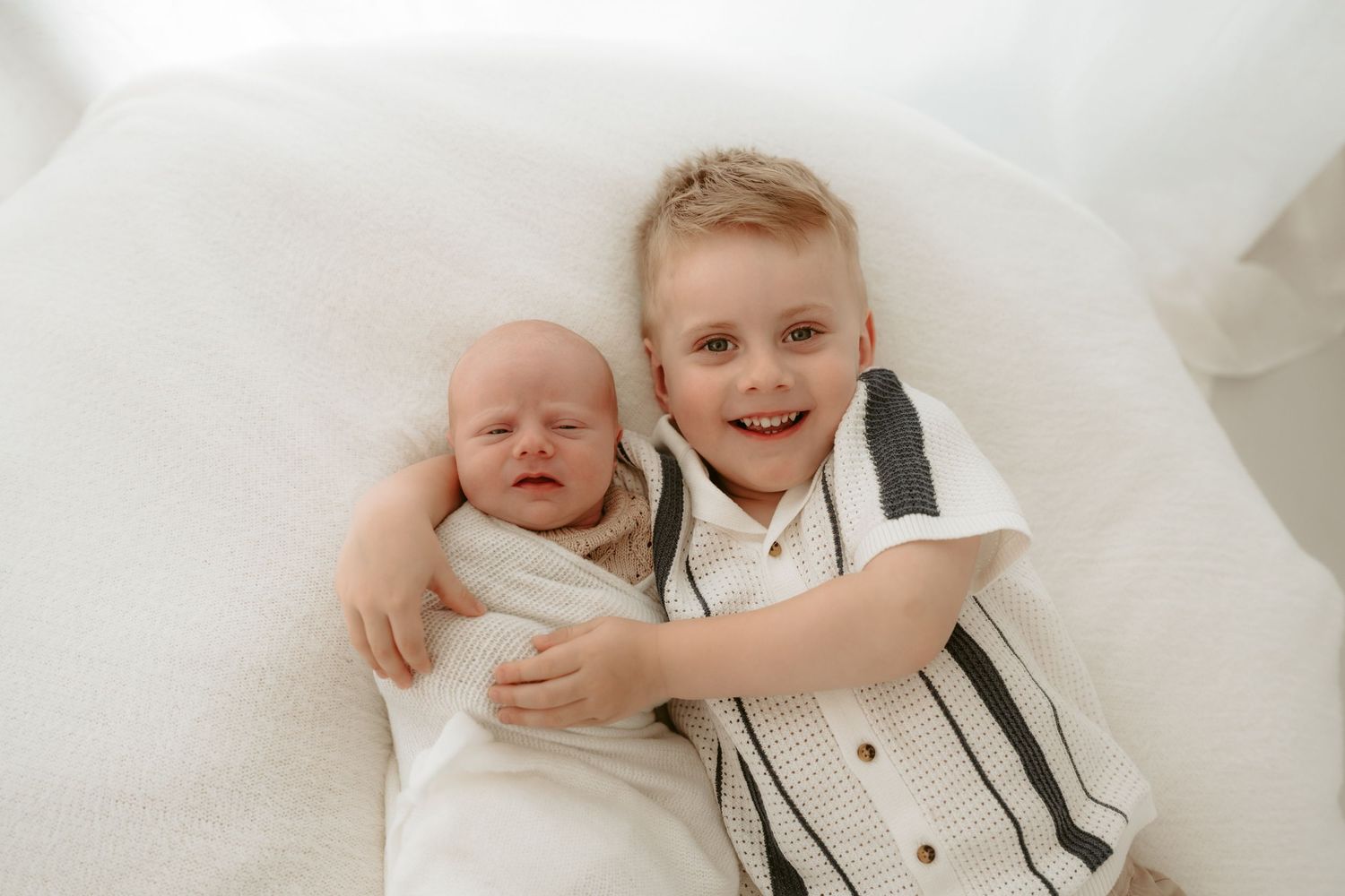 Two siblings bonding on white bedding with the older child embracing the baby.