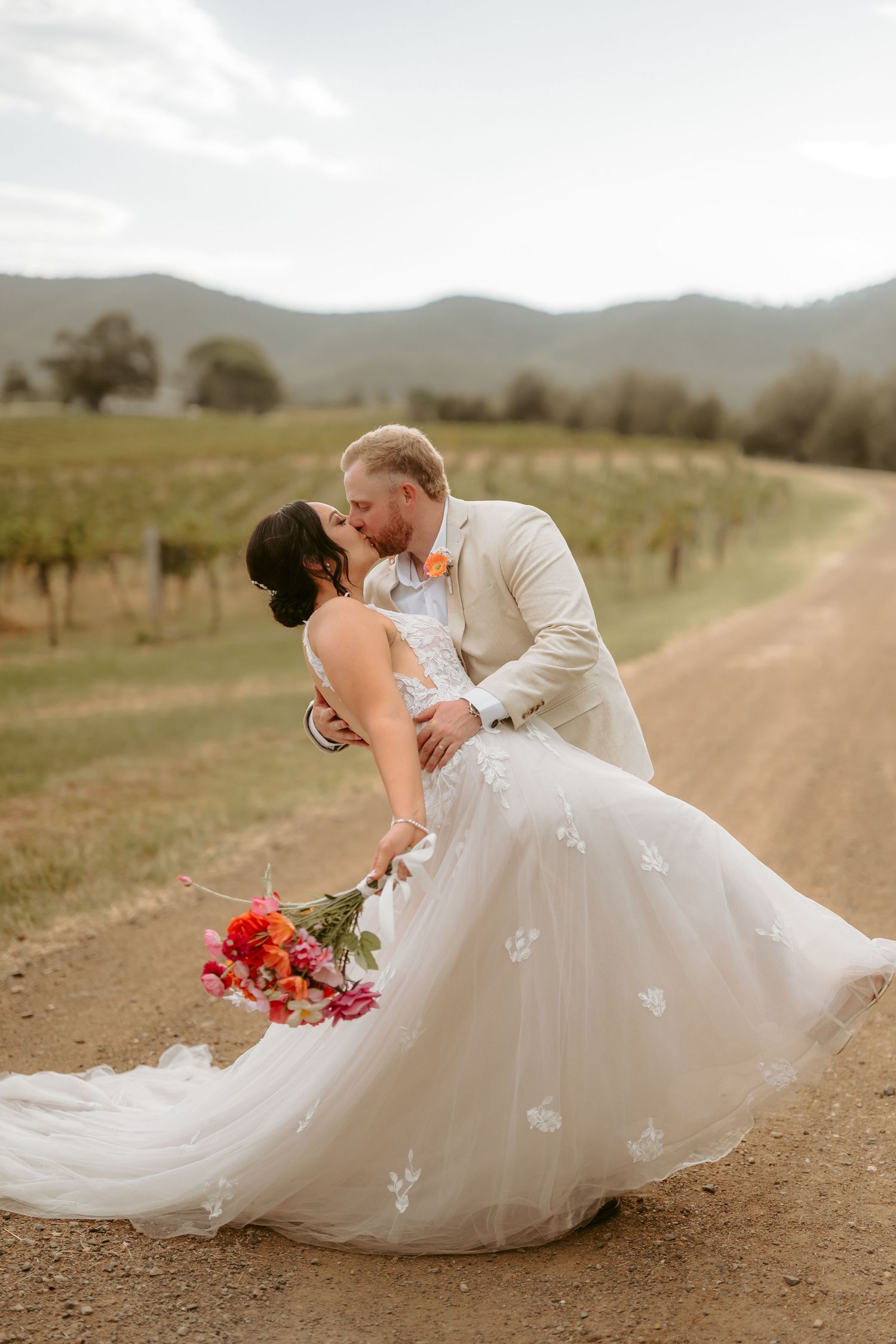 Romantic wedding dip pose with bride in flowing white dress against vineyard landscape.