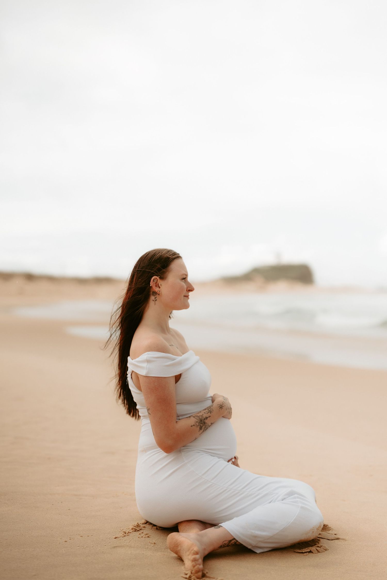 A pregnant woman in white dress poses joyfully on beach during sunset.